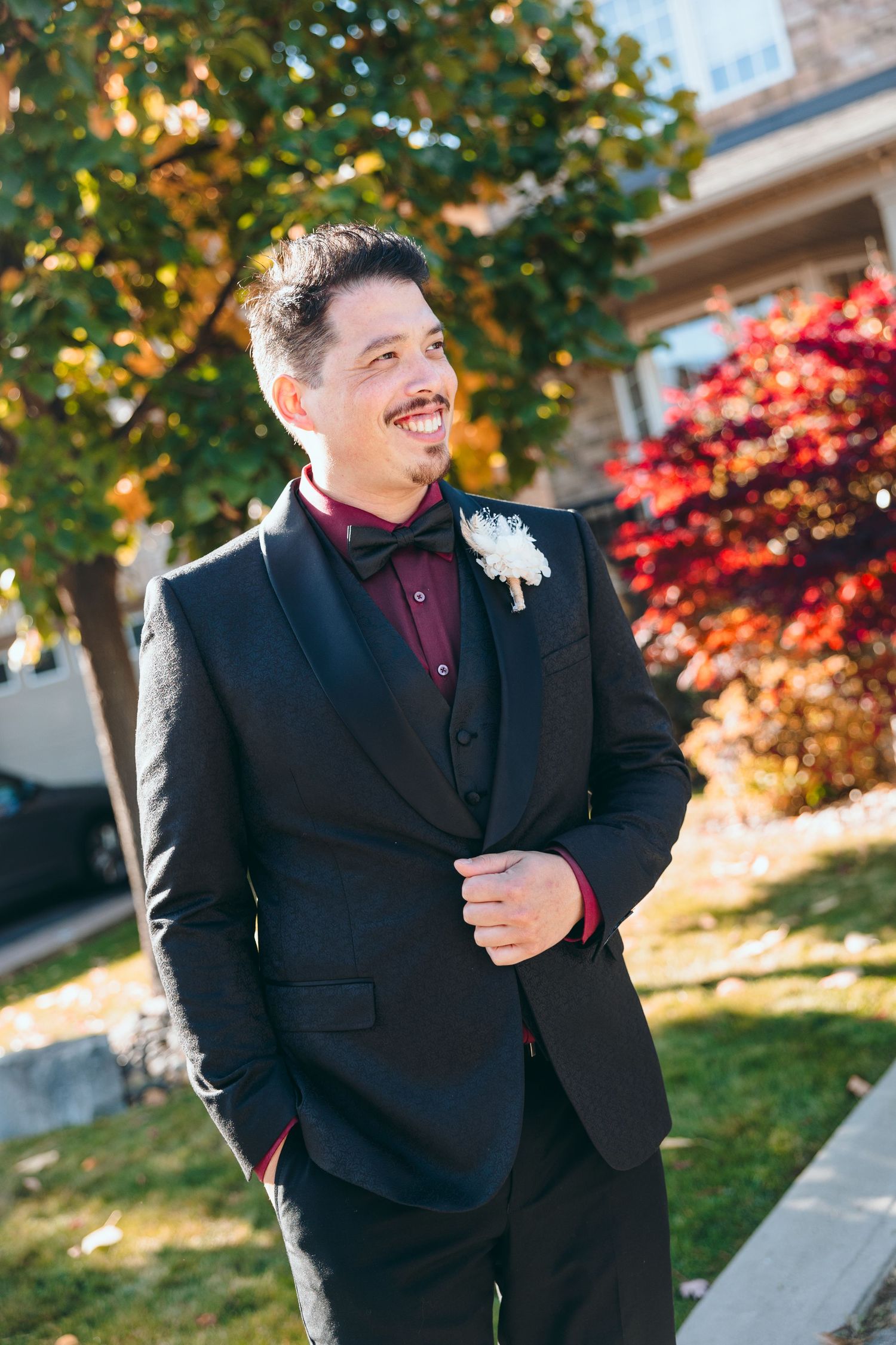 Person in black suit and burgundy tie poses outdoors against fall foliage.