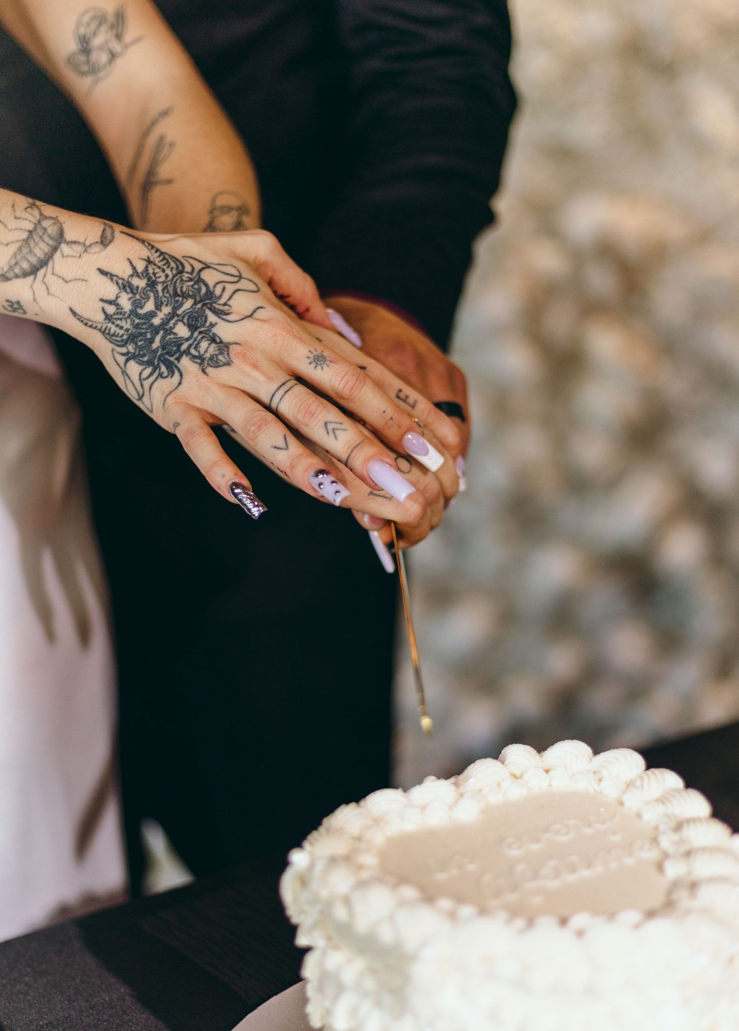 Tattooed hands cutting into a white wedding cake with delicate details.