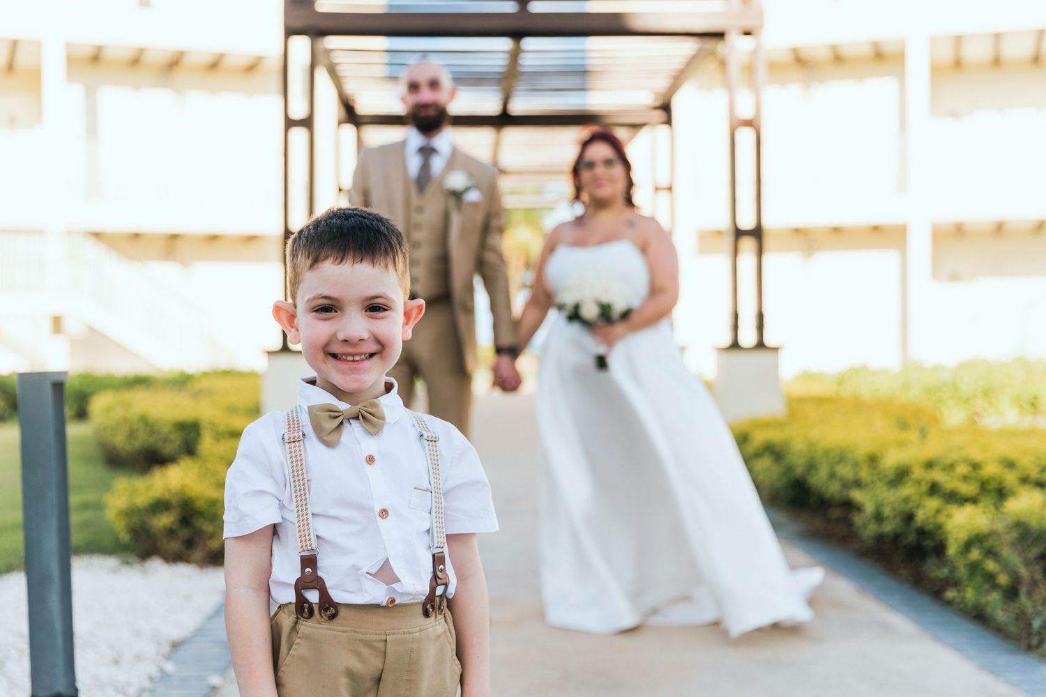 A young ring bearer in suspenders smiles at the camera during an outdoor wedding ceremony.