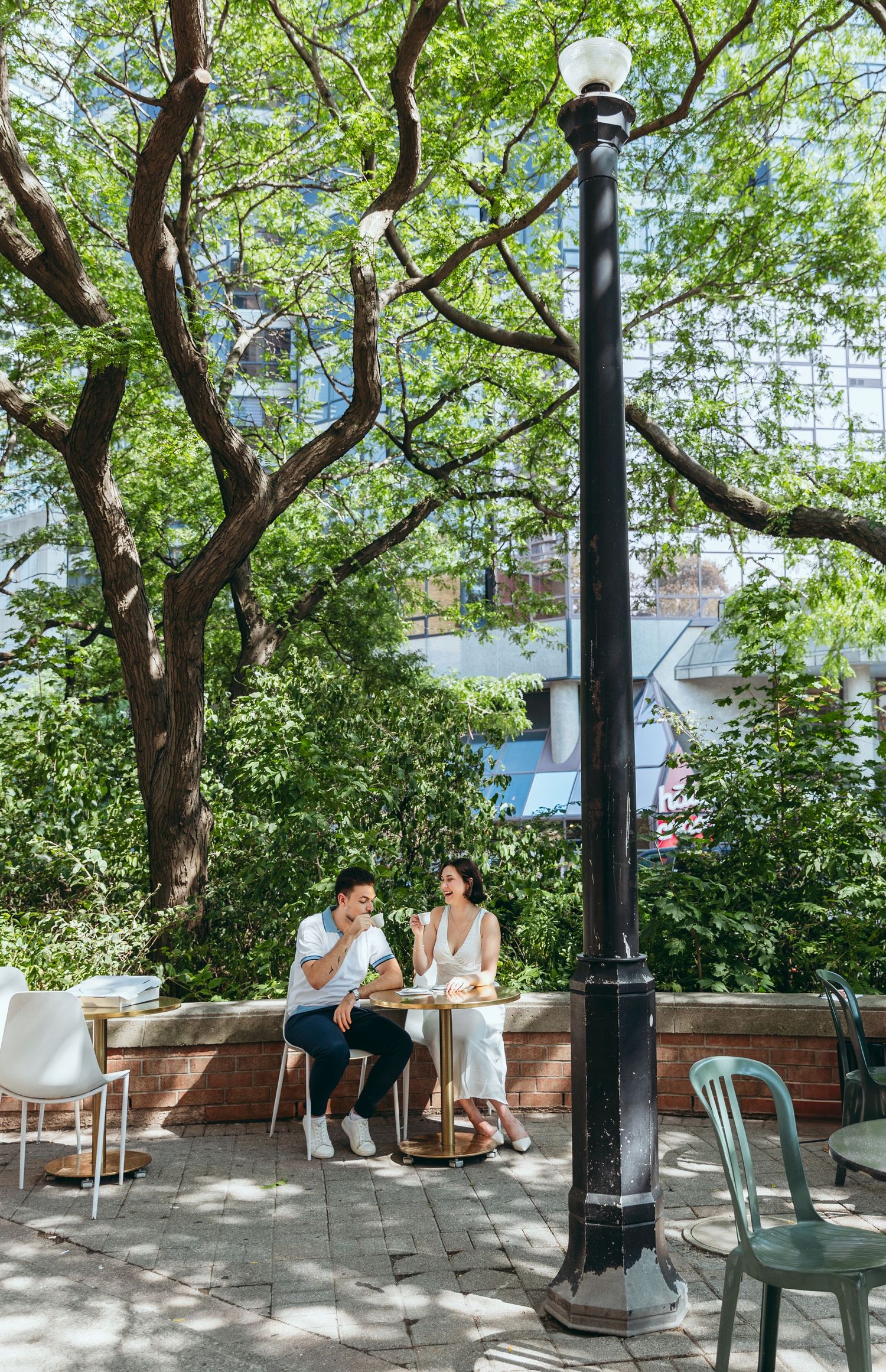 A couple enjoys coffee at an outdoor cafe table under green trees on a sunny day.