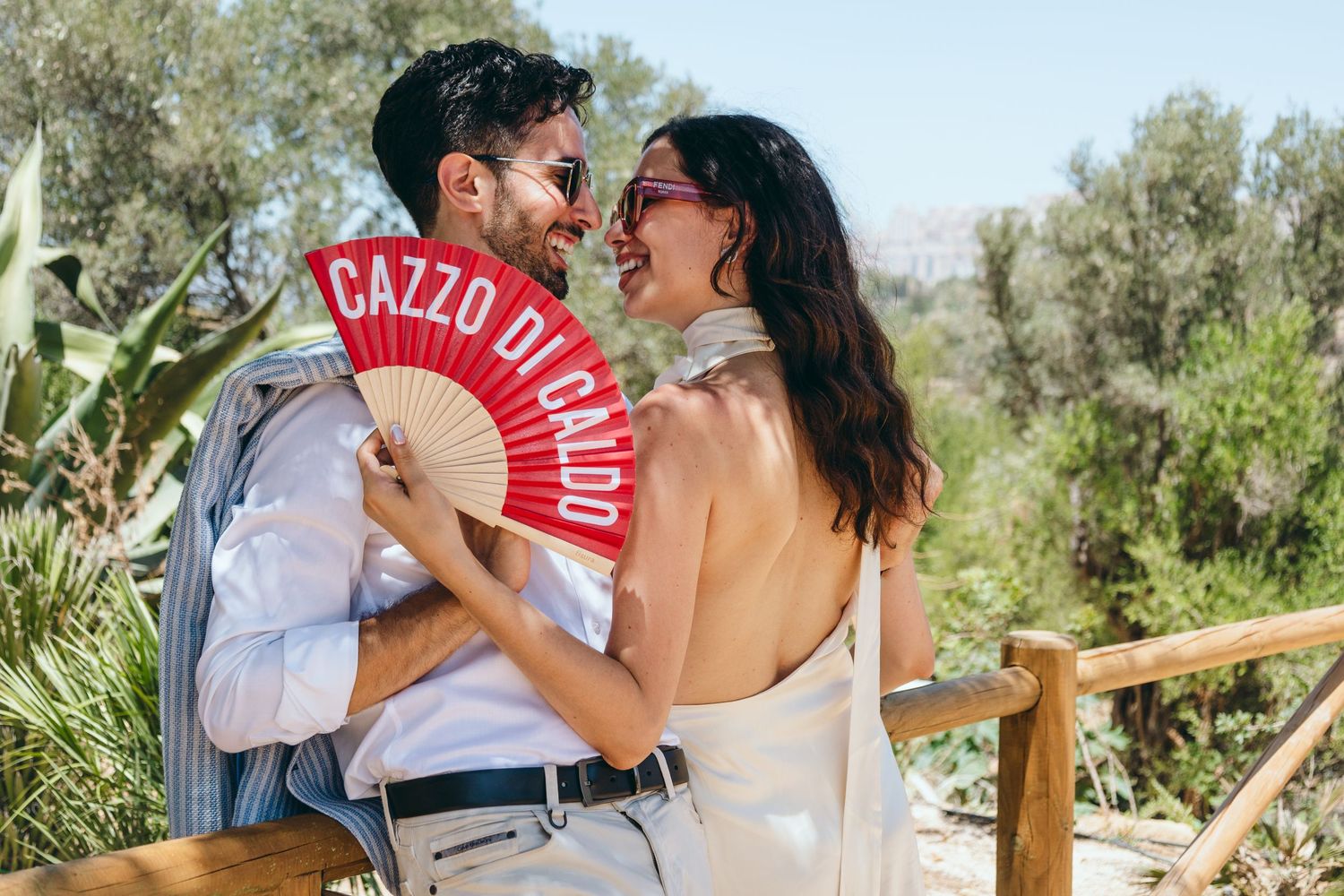 Playful couple in white outfits pose with red fan against natural garden backdrop.