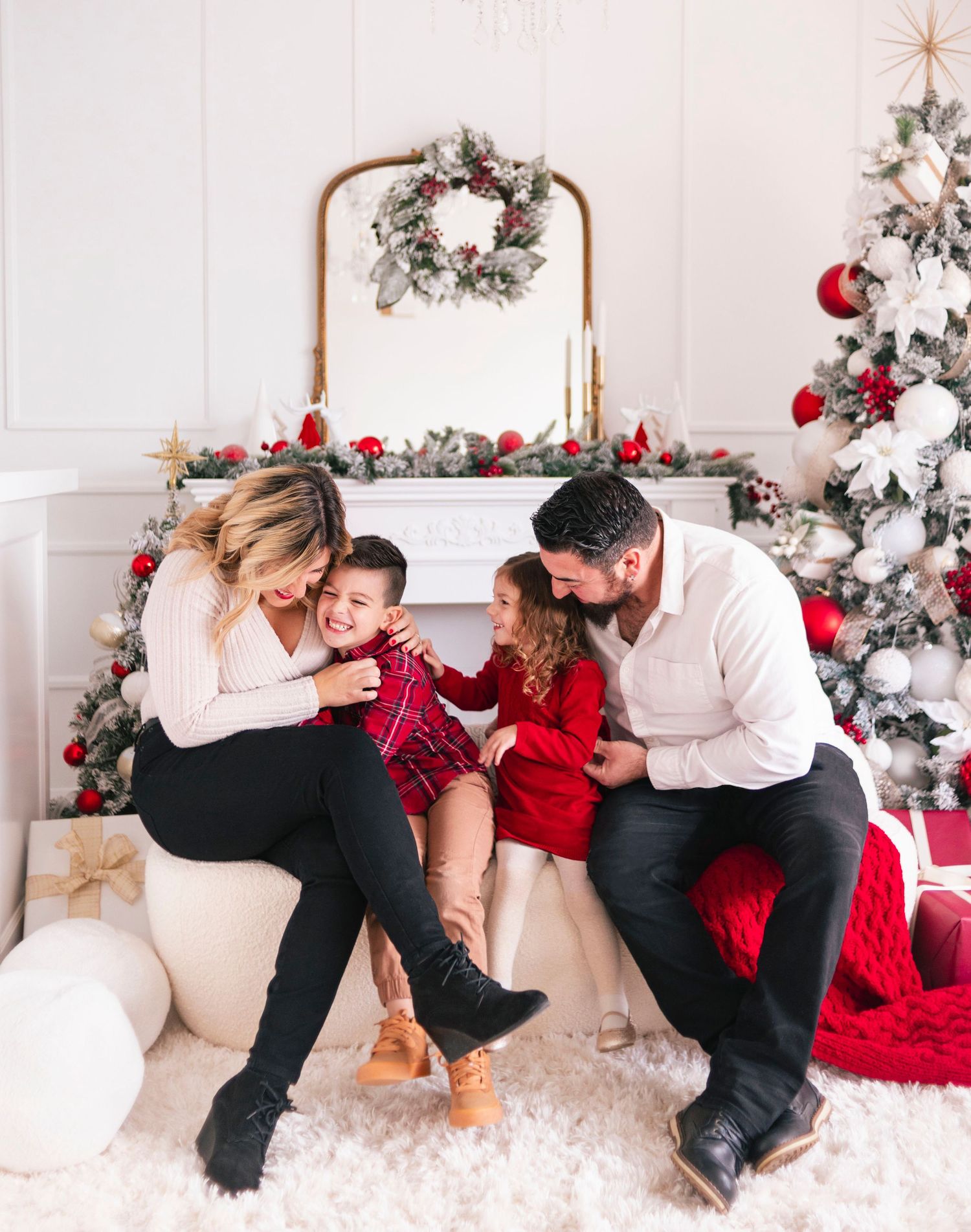 Family of four sharing joyful Christmas moments by a decorated fireplace and festive tree in a bright white holiday setting.