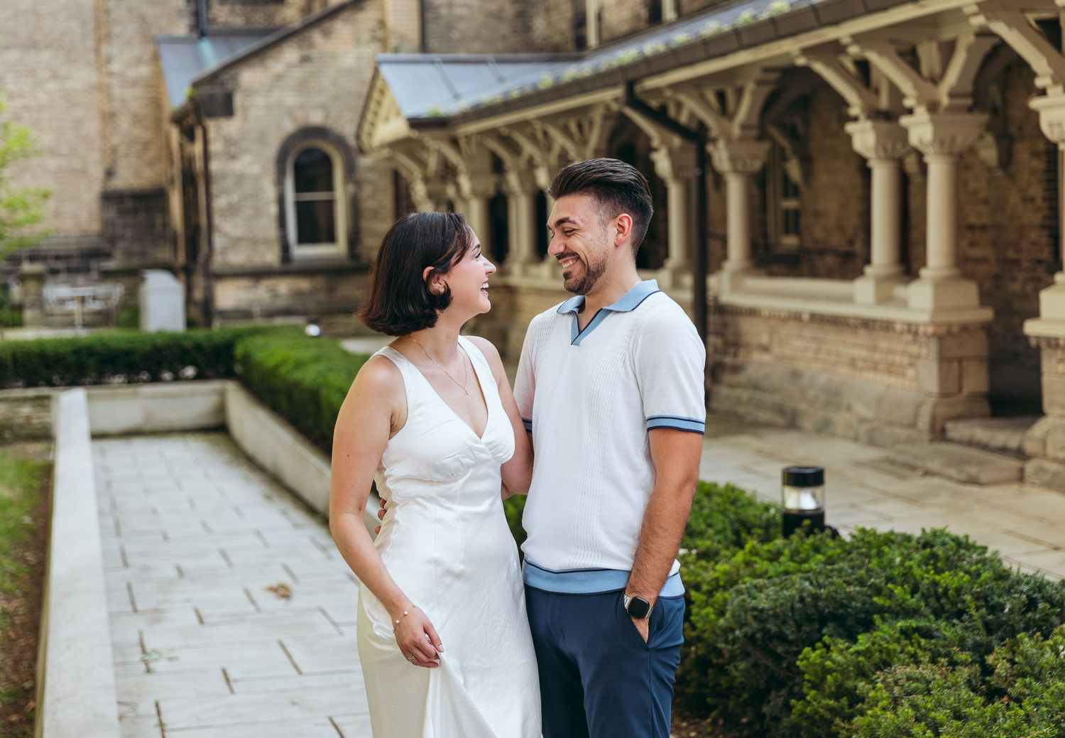 A couple shares an intimate moment along a historic stone colonnade with manicured hedges.