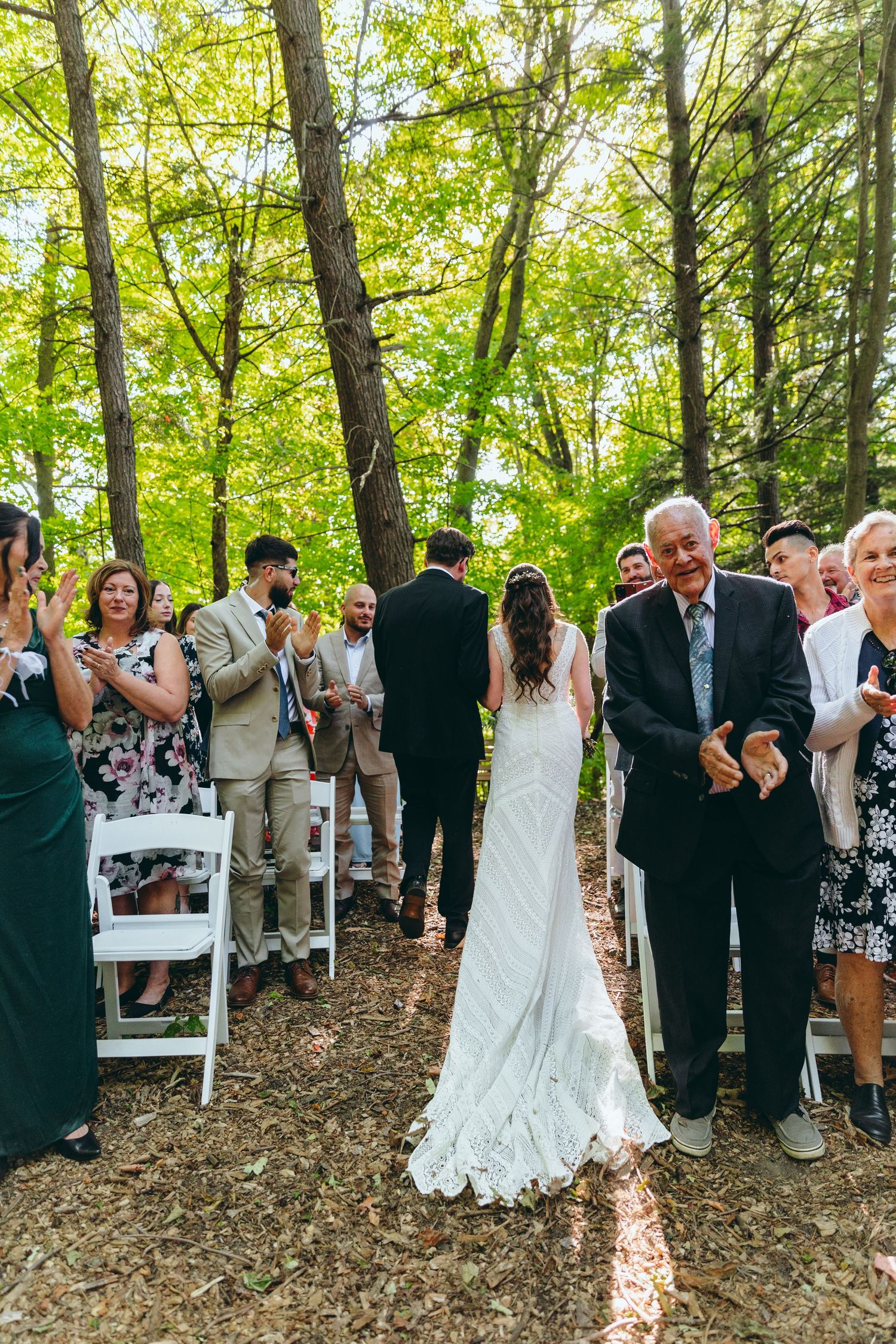 Wedding guests celebrate during a ceremony in a lush green forest setting with white chairs.