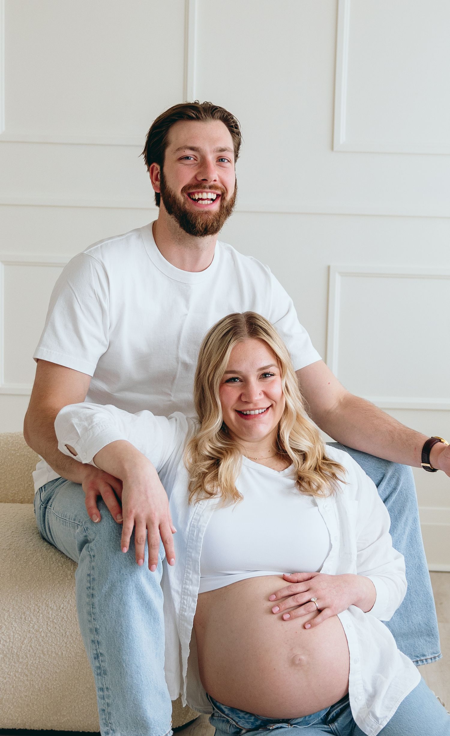 Couple in white shirts and jeans sitting together on floor during maternity photoshoot.