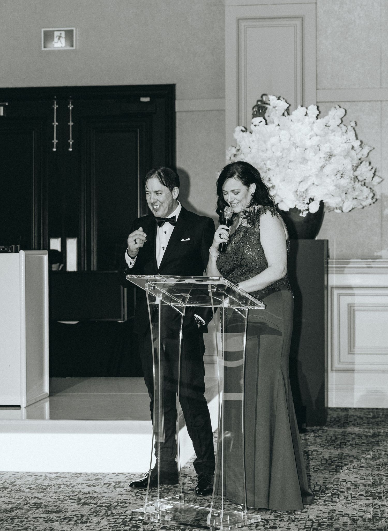 Two speakers stand at clear podium with white floral arrangement in formal event setting.
