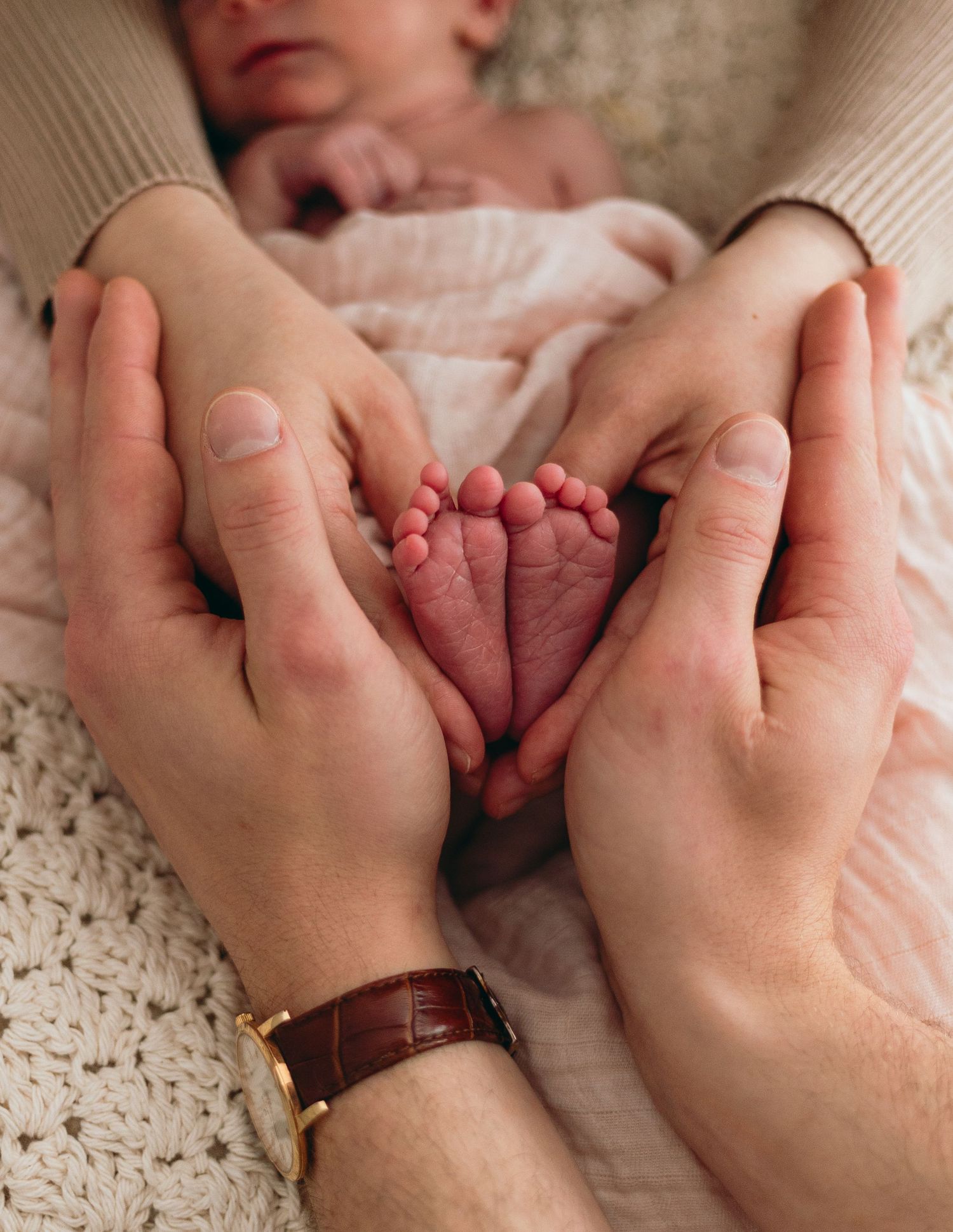 Close-up view of tiny newborn baby feet cradled gently in adult hands on a textured white blanket background.