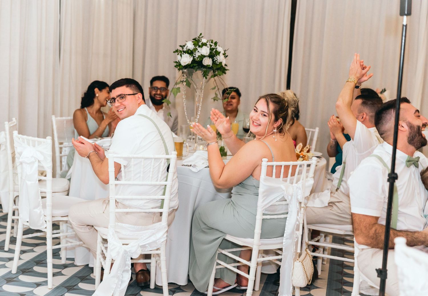 Wedding guests seated at white tables celebrate during reception festivities.