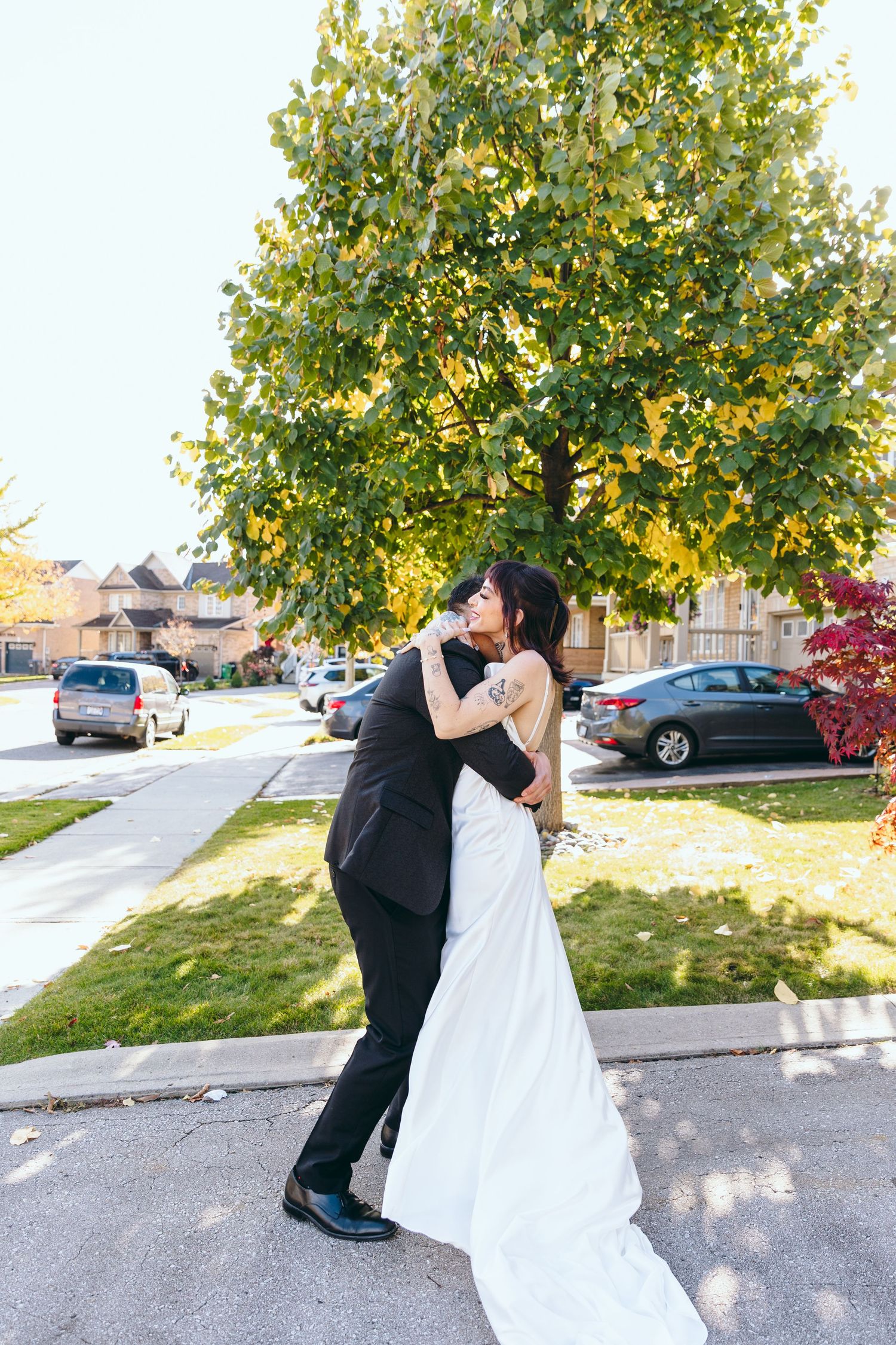 A couple embracing in their wedding attire on a suburban street with fall trees.