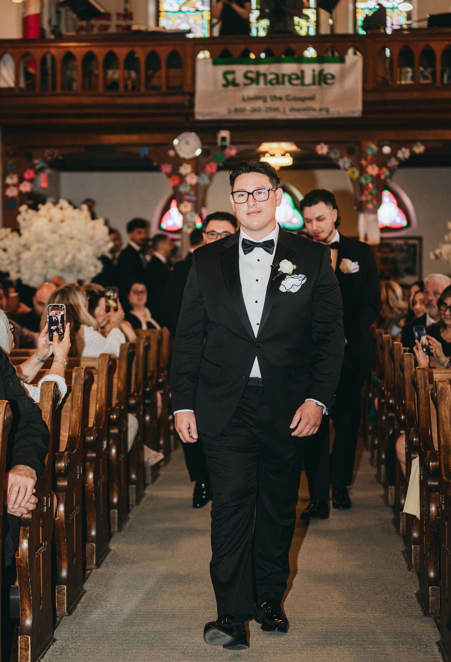 A groomsman in a black tuxedo and bow tie walks down the church aisle during a wedding ceremony.
