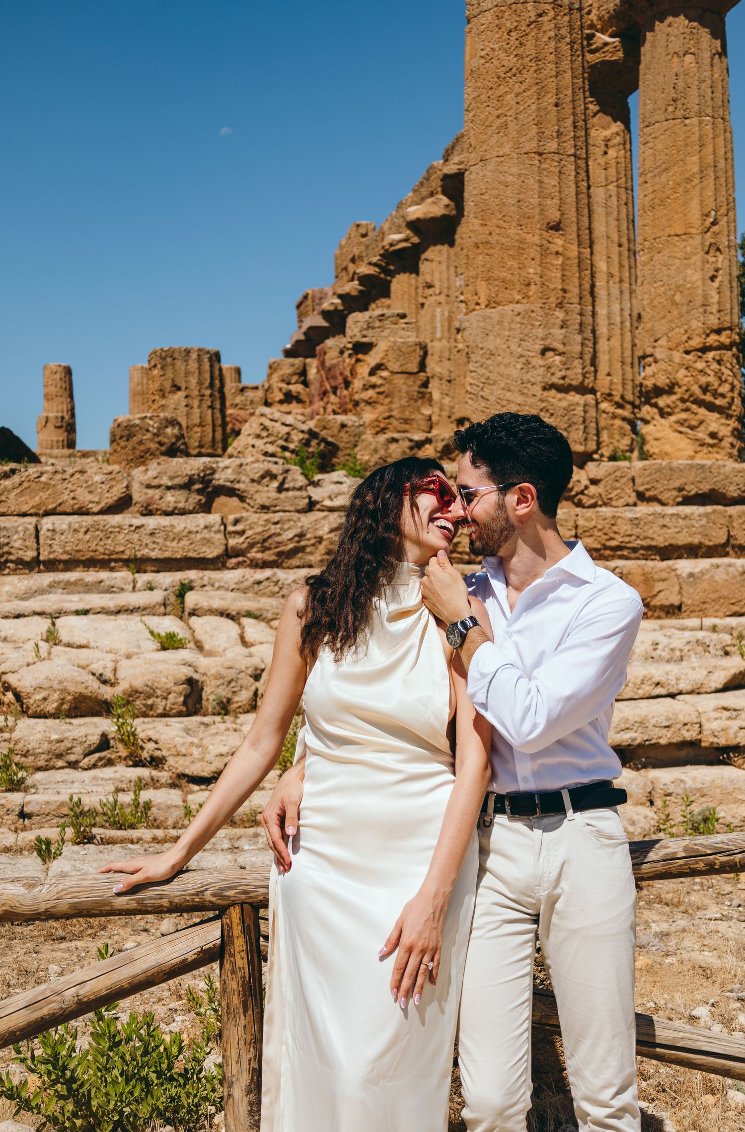 A romantic photoshoot at ancient Greek ruins with a couple in white outfits posing against stone columns.