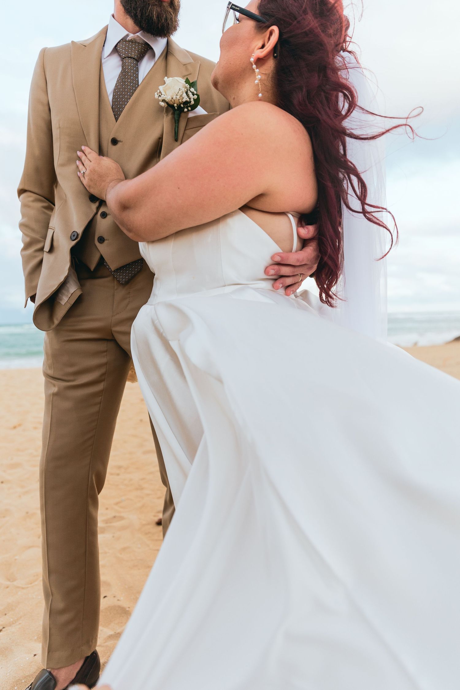 A couple in wedding attire embrace on a beach with flowing dress and tan suit.