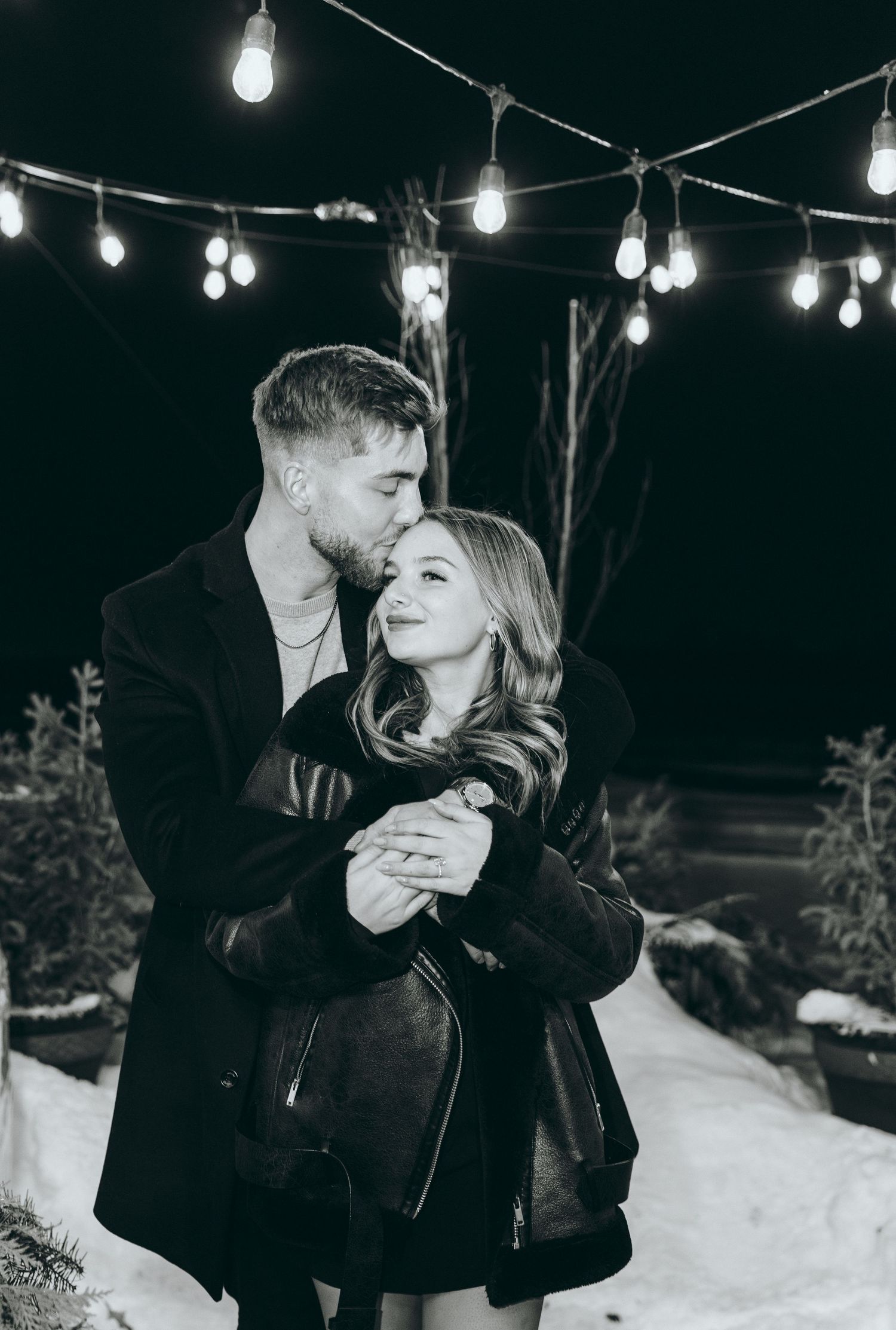 Black and white photo of couple under string lights in snowy setting.