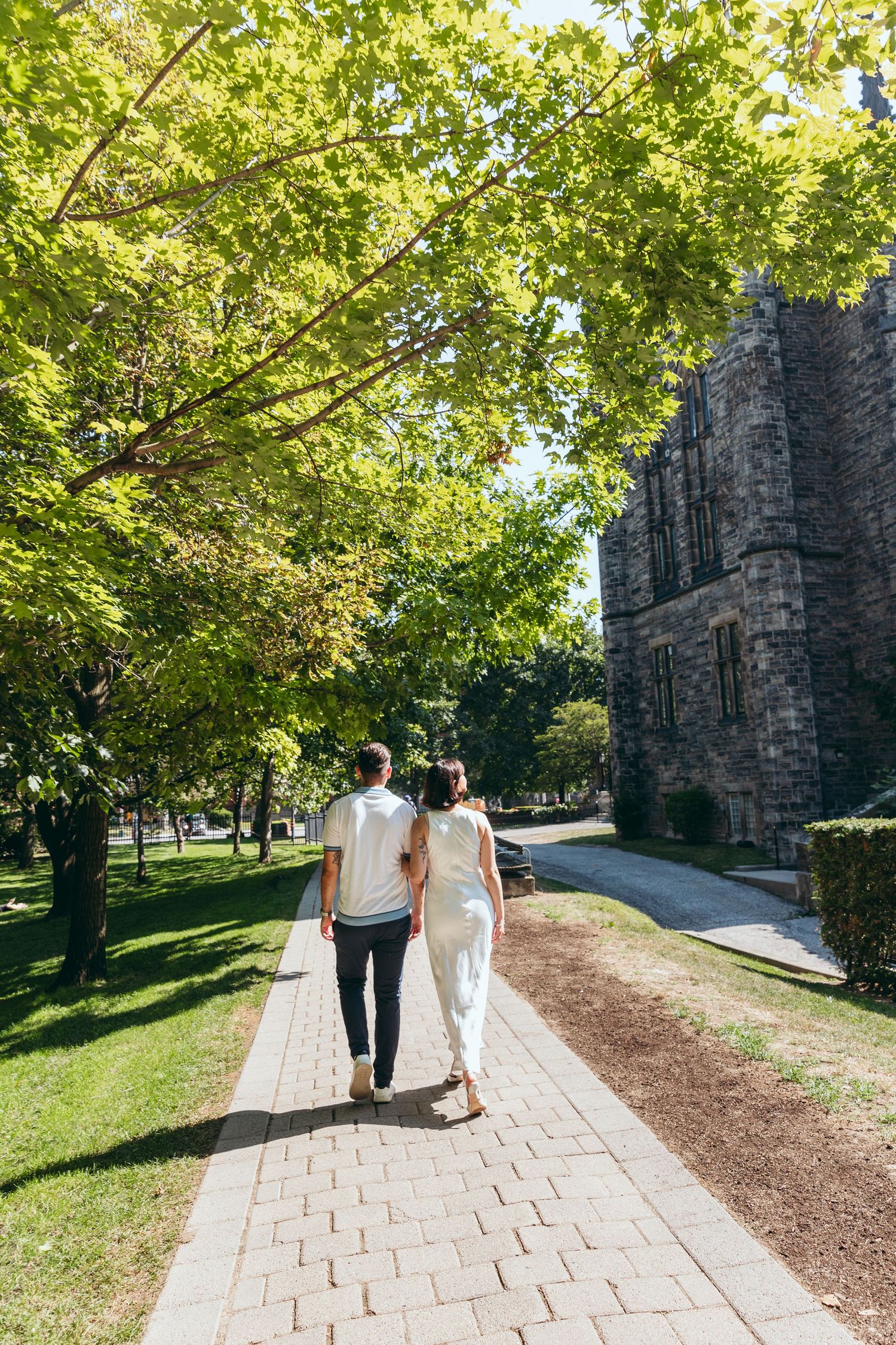 A couple walks along a stone path beside a Gothic-style university building on a sunny day.