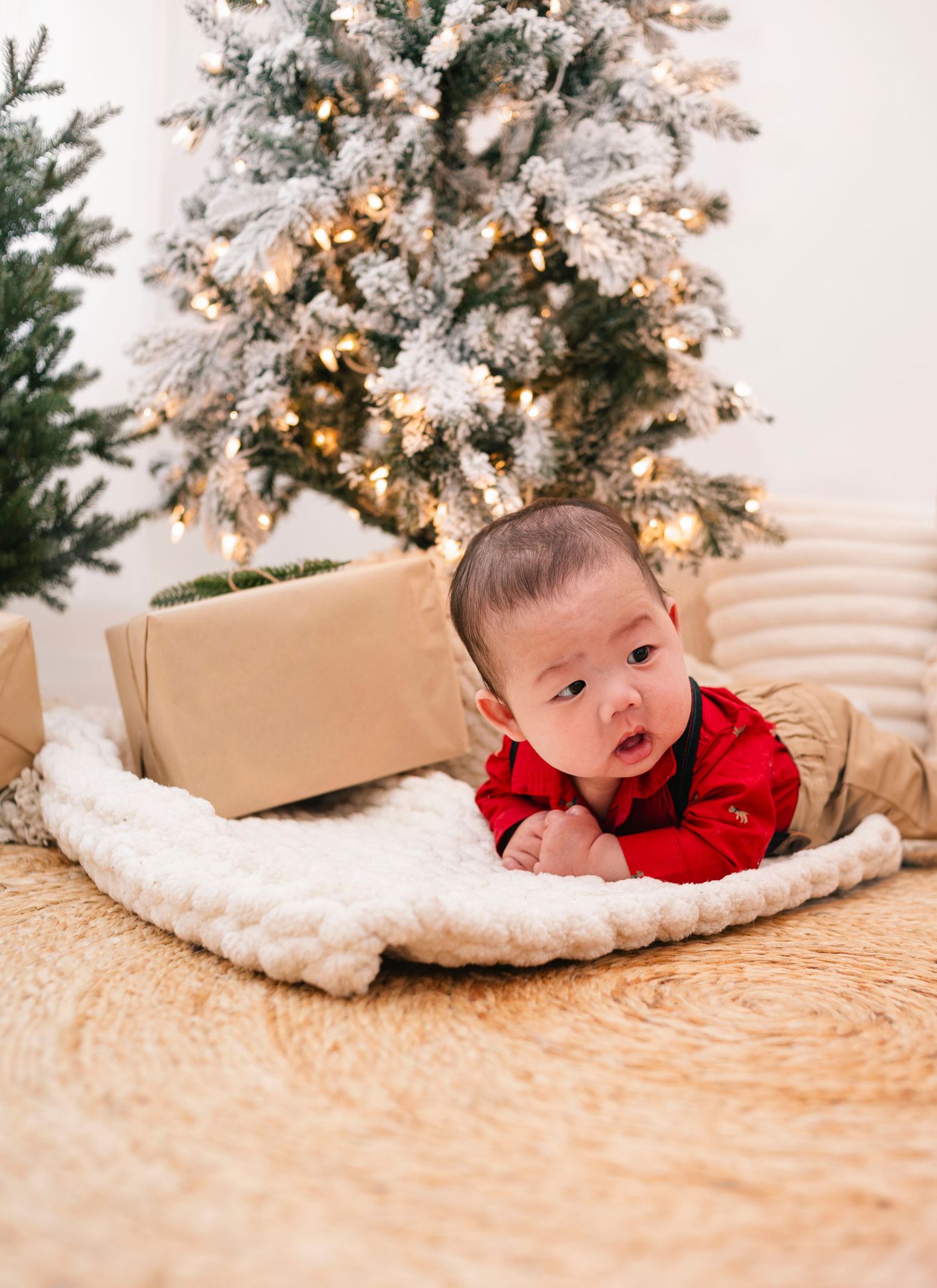 Baby in red outfit doing tummy time on fluffy blanket in front of a snow-flocked Christmas tree with warm lights.