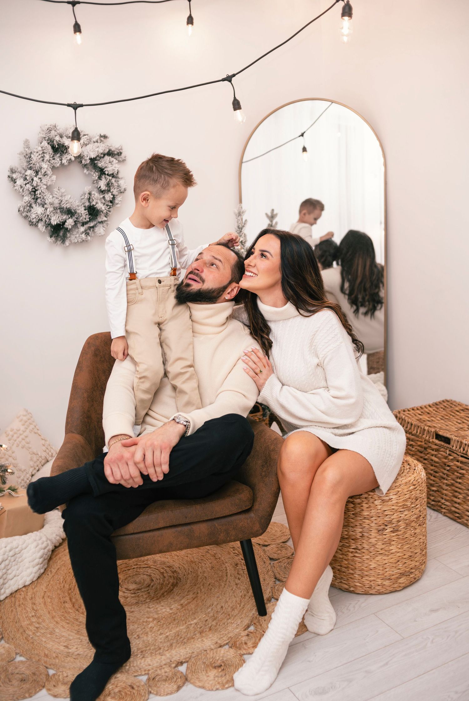 Family with young son enjoying a cozy Christmas photo session in a neutral-toned studio with string lights.