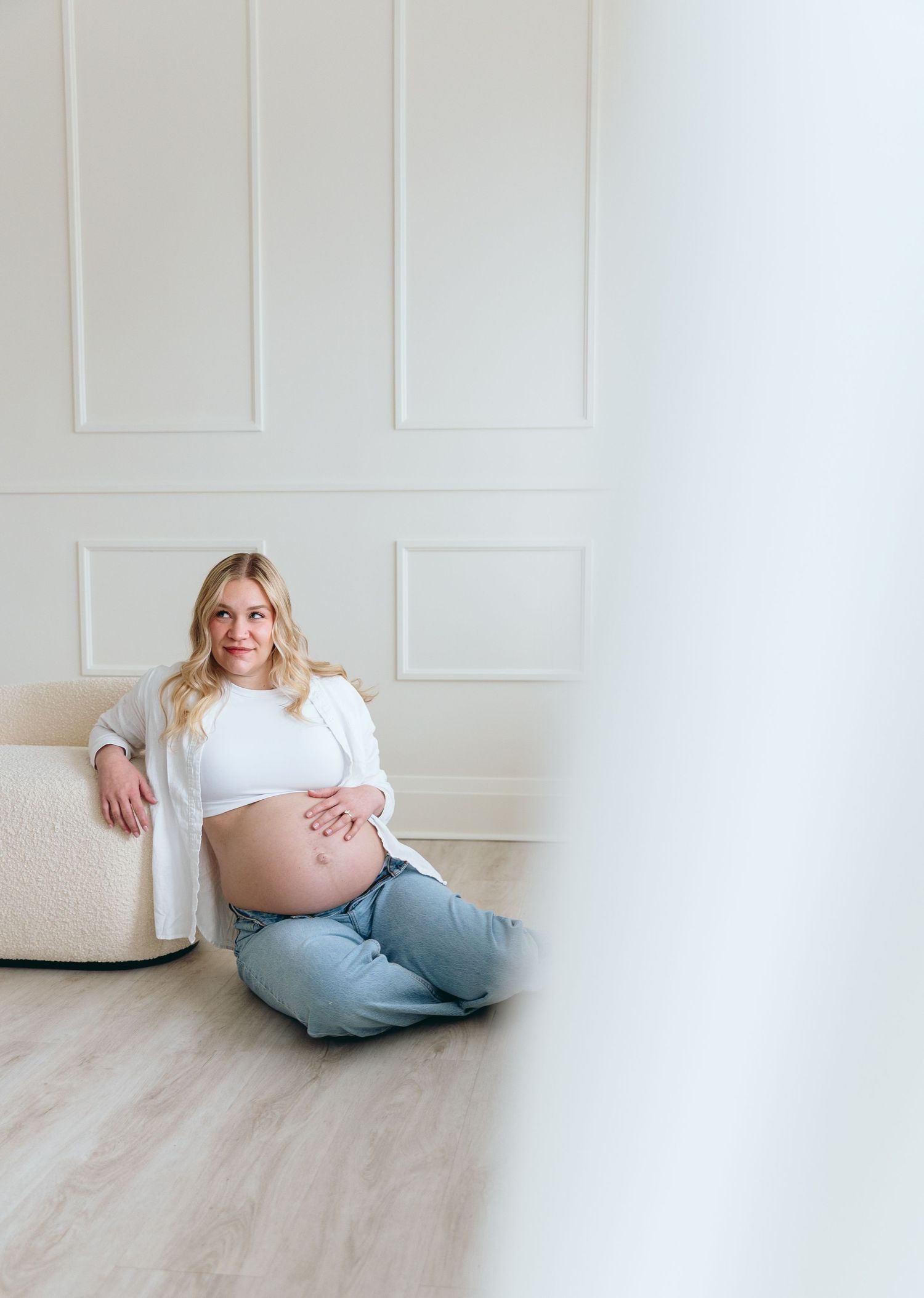 Expectant mother relaxing on floor against white paneled wall in casual attire.