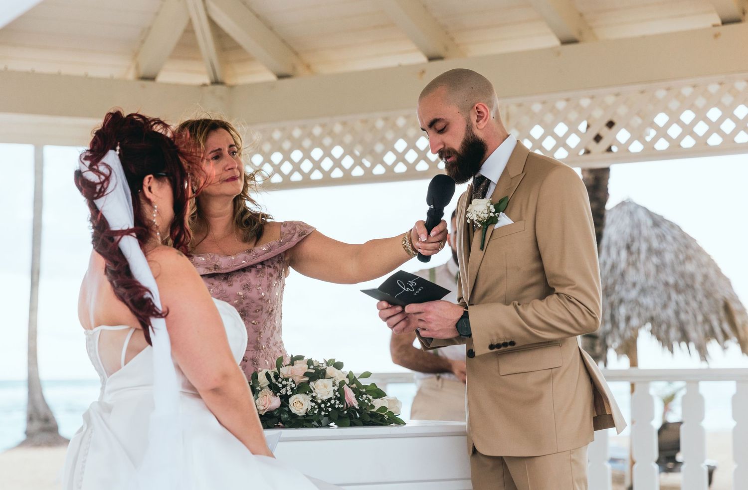 Wedding guests gather under a beachside gazebo with palm trees in the background.