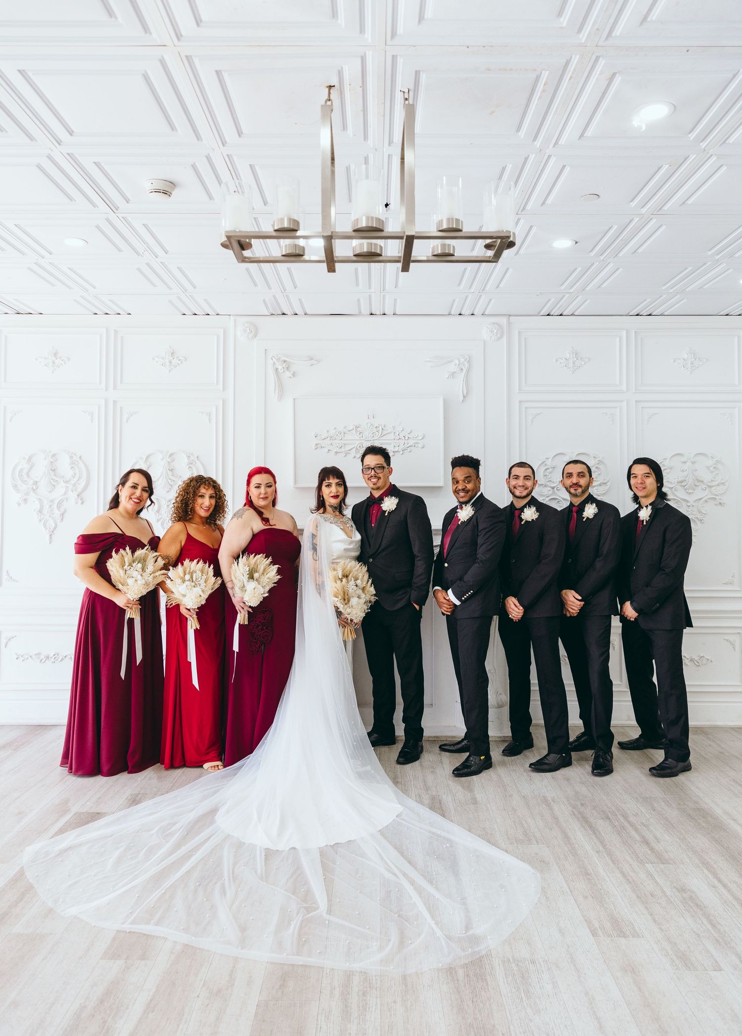 Wedding party in burgundy dresses and black suits posing in an elegant white room.