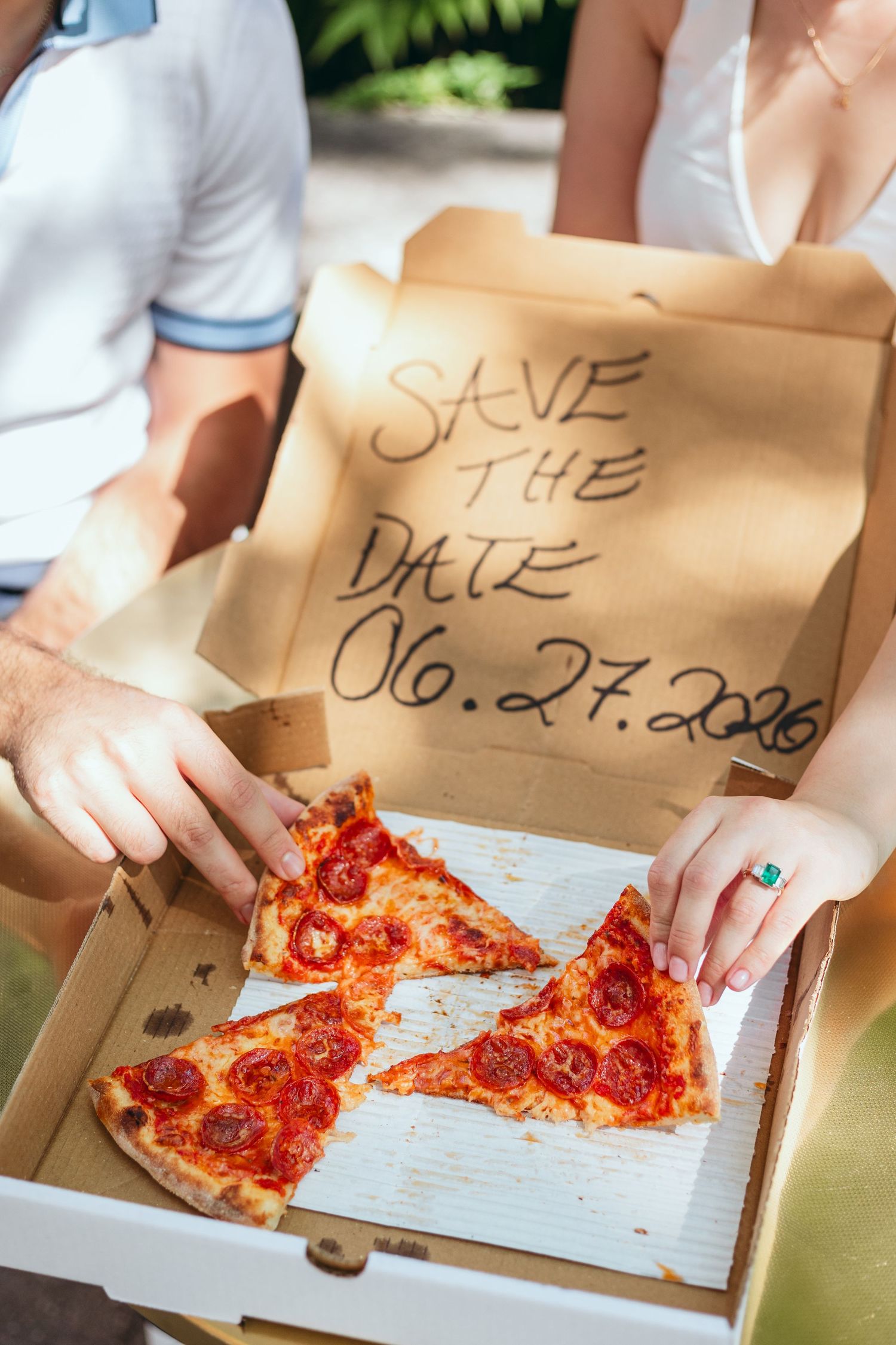 Close-up of hands holding pizza slices with a save the date note visible in the box.