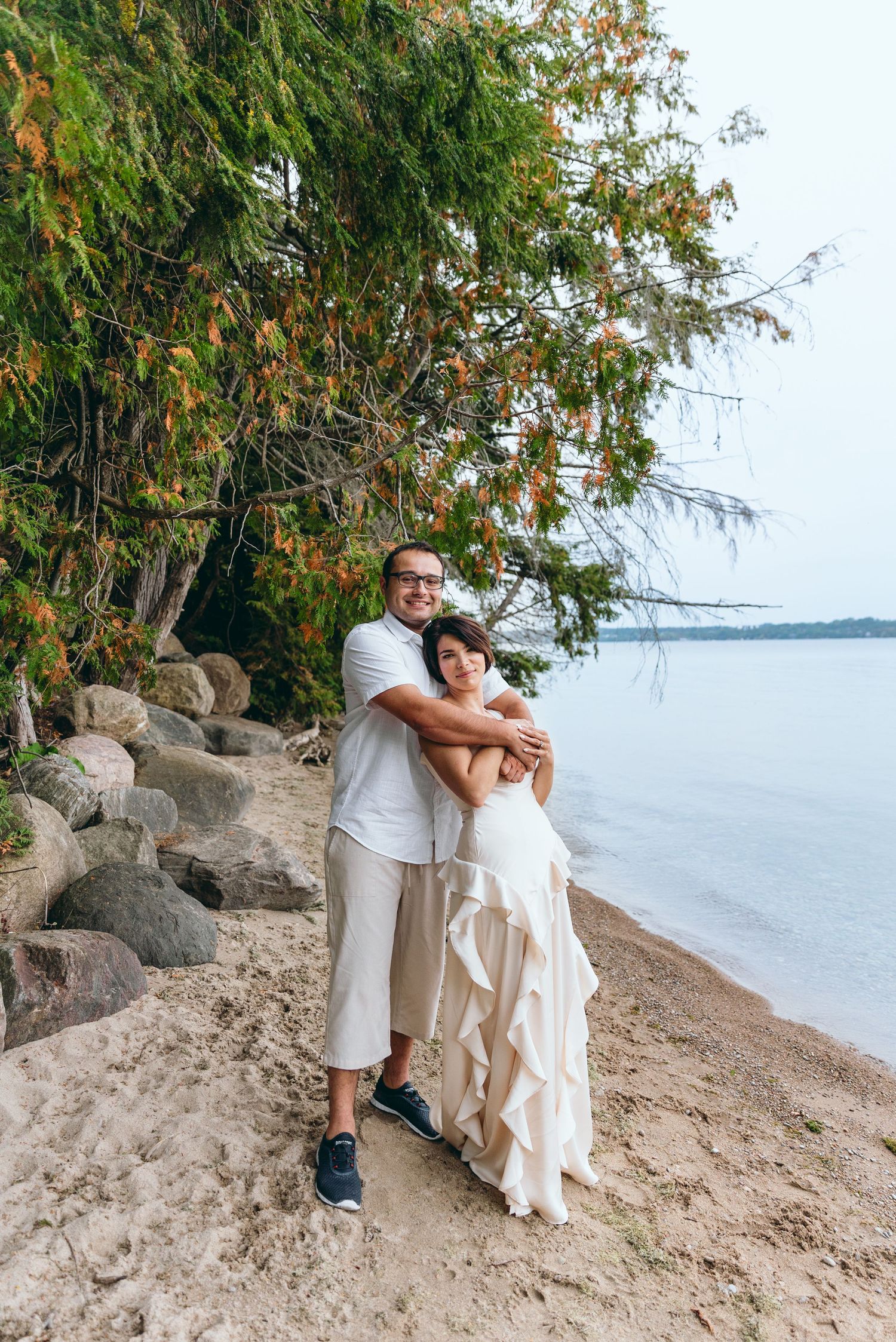 A couple in white clothing hugging on a beach with rocks and trees behind them.