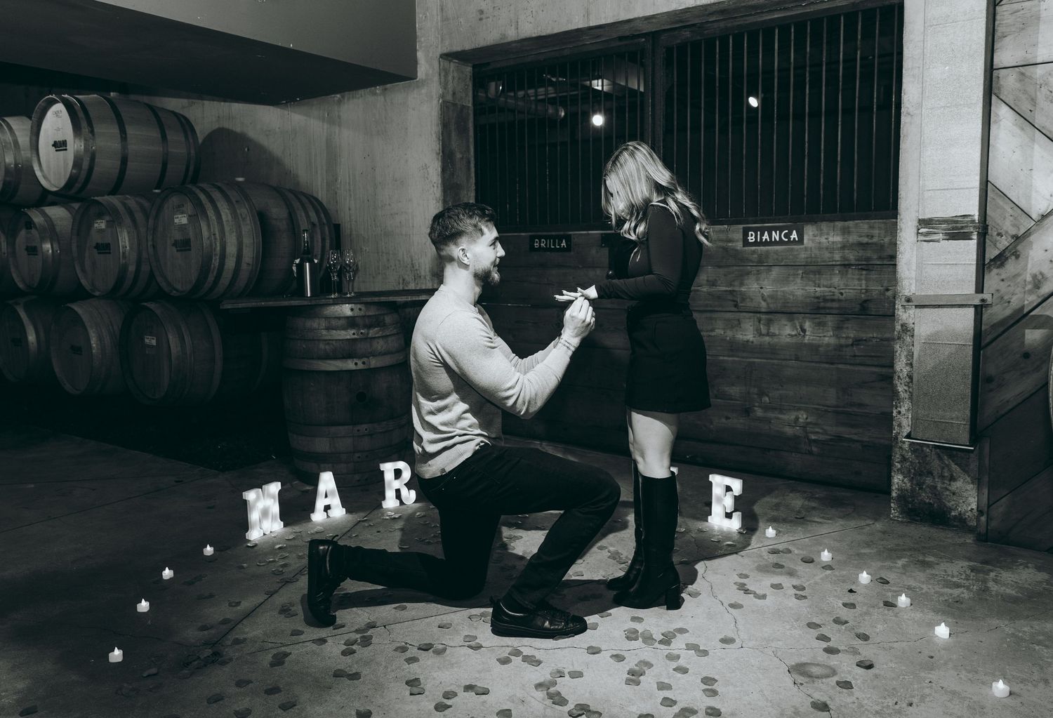 Indoor engagement scene with white letters on floor and wooden barrels in background.