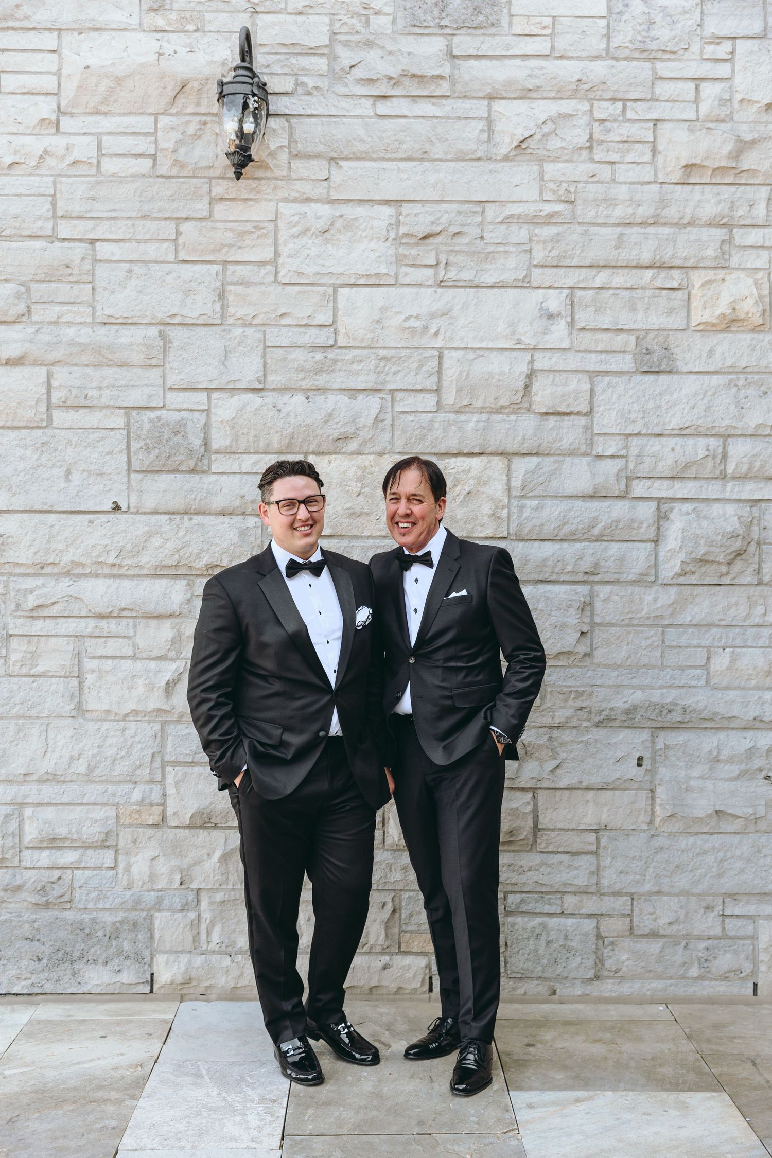 Two people in black tuxedos standing against stone wall.