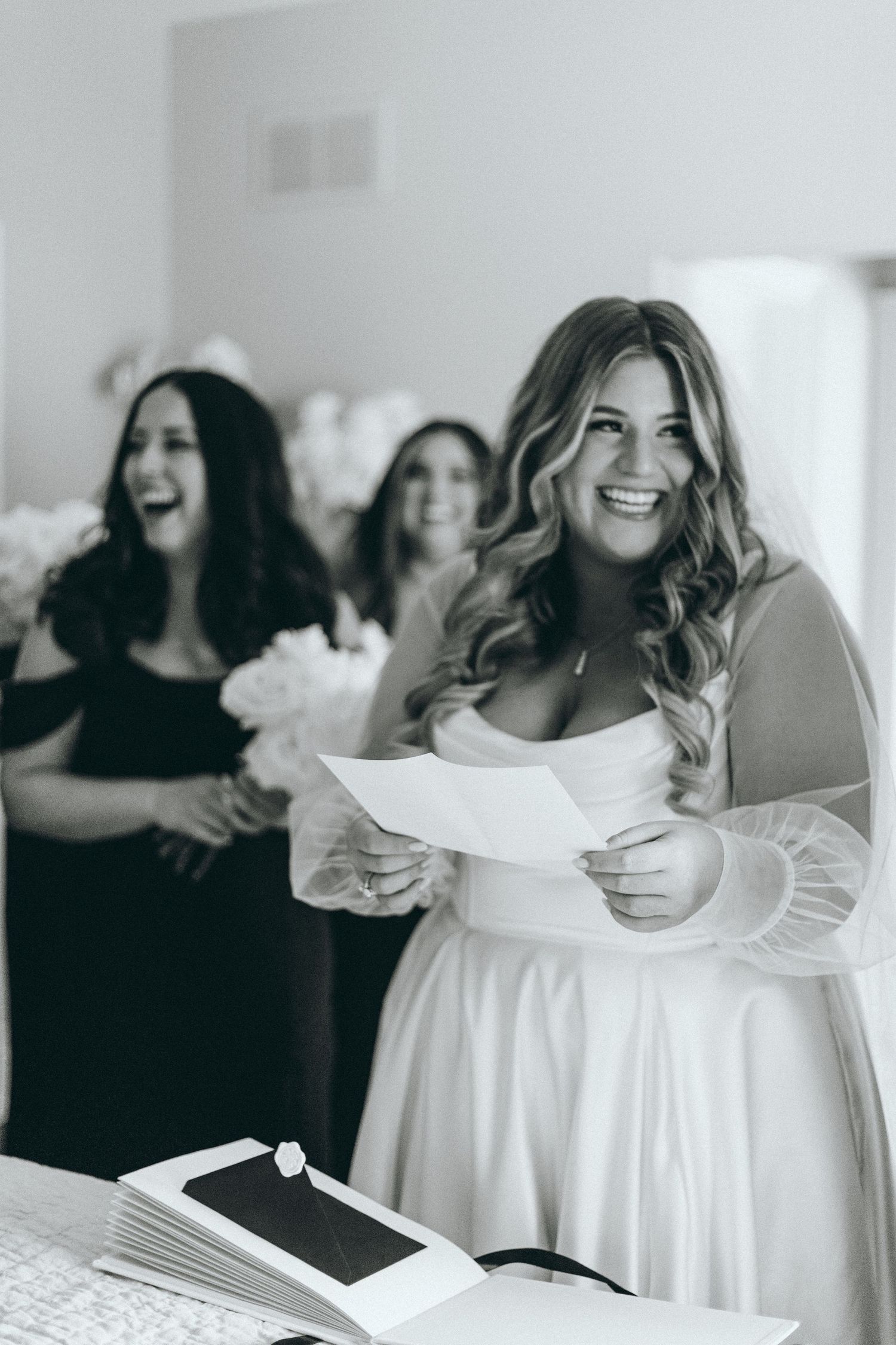 Bridesmaids in white dresses share joyful moment while reading together.
