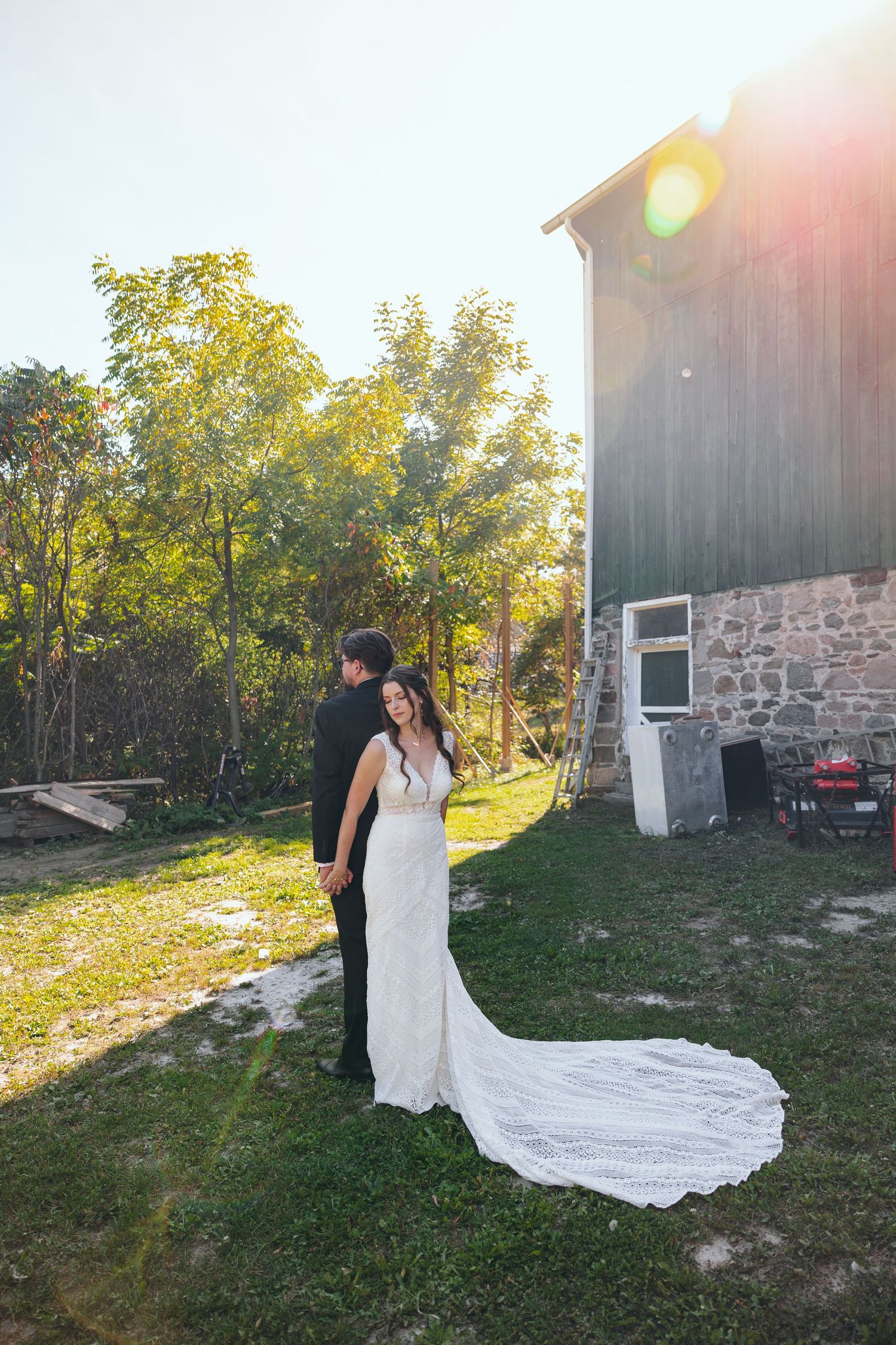 Wedding couple poses near rustic barn with flowing white dress train in golden evening light.