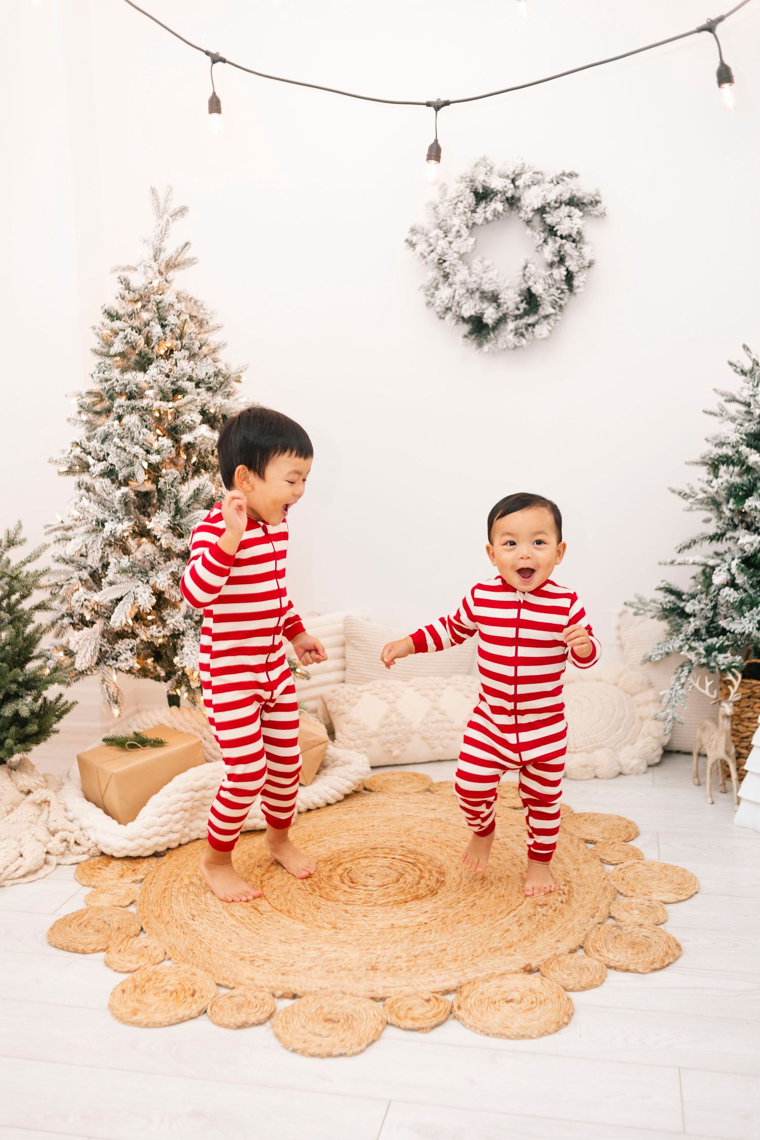 Two young children in matching red and white striped Christmas pajamas playing near decorated holiday trees.