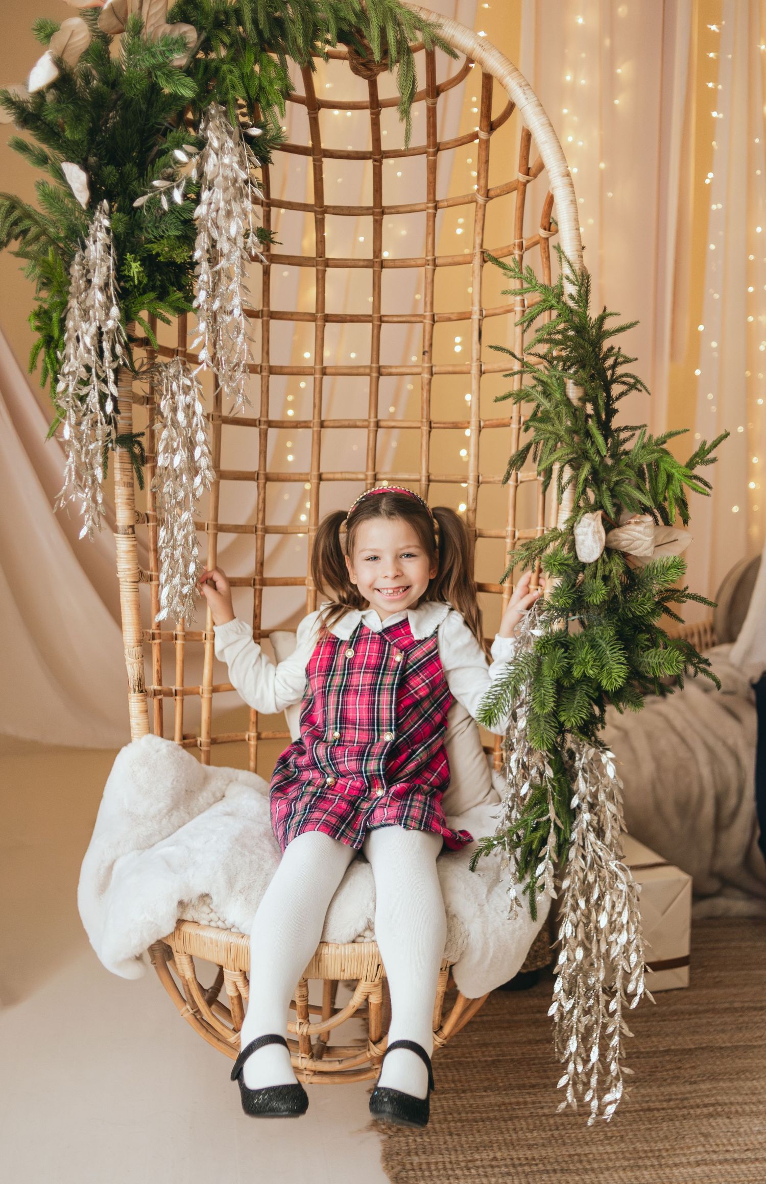 A smiling girl in a plaid dress sits in a decorated wicker swing chair with Christmas greenery and fairy lights.