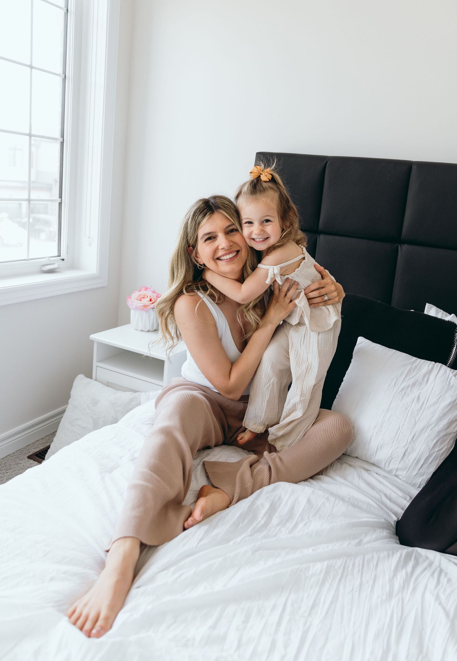 A tender family moment captured on a white bed with black headboard near a bright window.