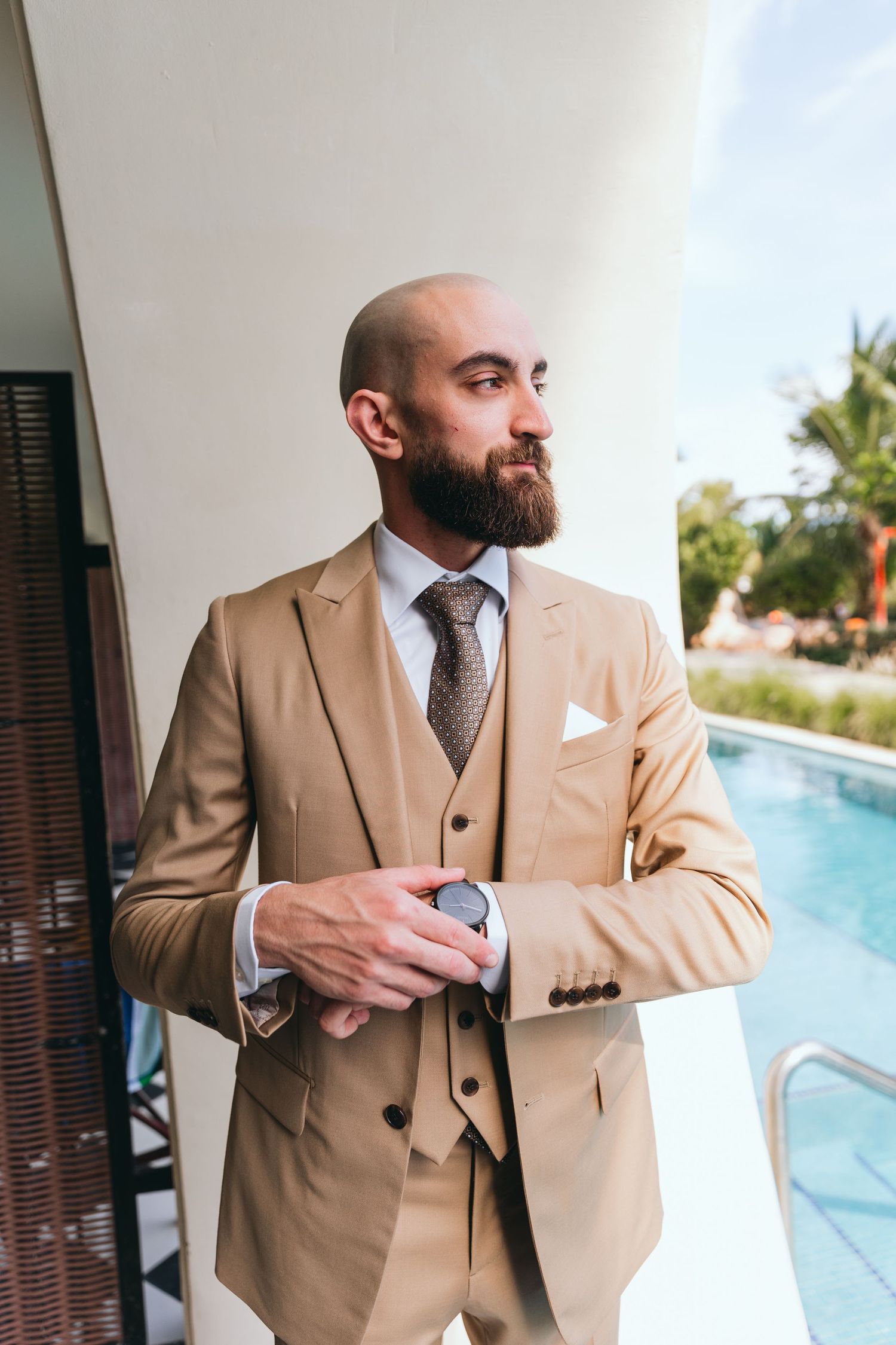 Groom in tan three-piece suit poses near a pool during a destination wedding photoshoot.