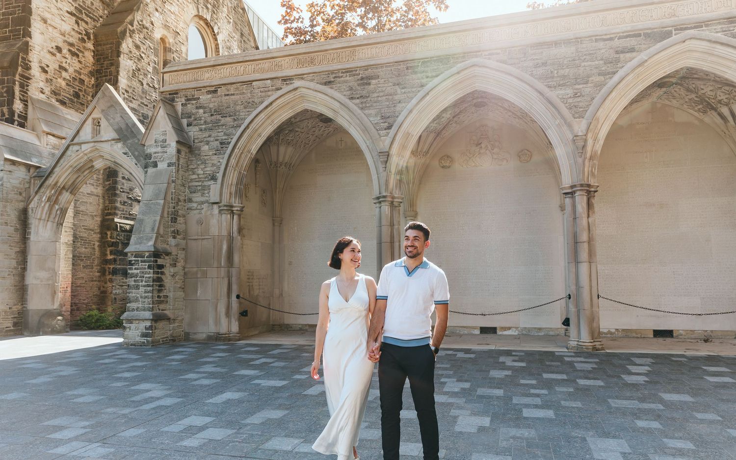 A couple stands together in a historic stone colonnade with gothic architectural details.
