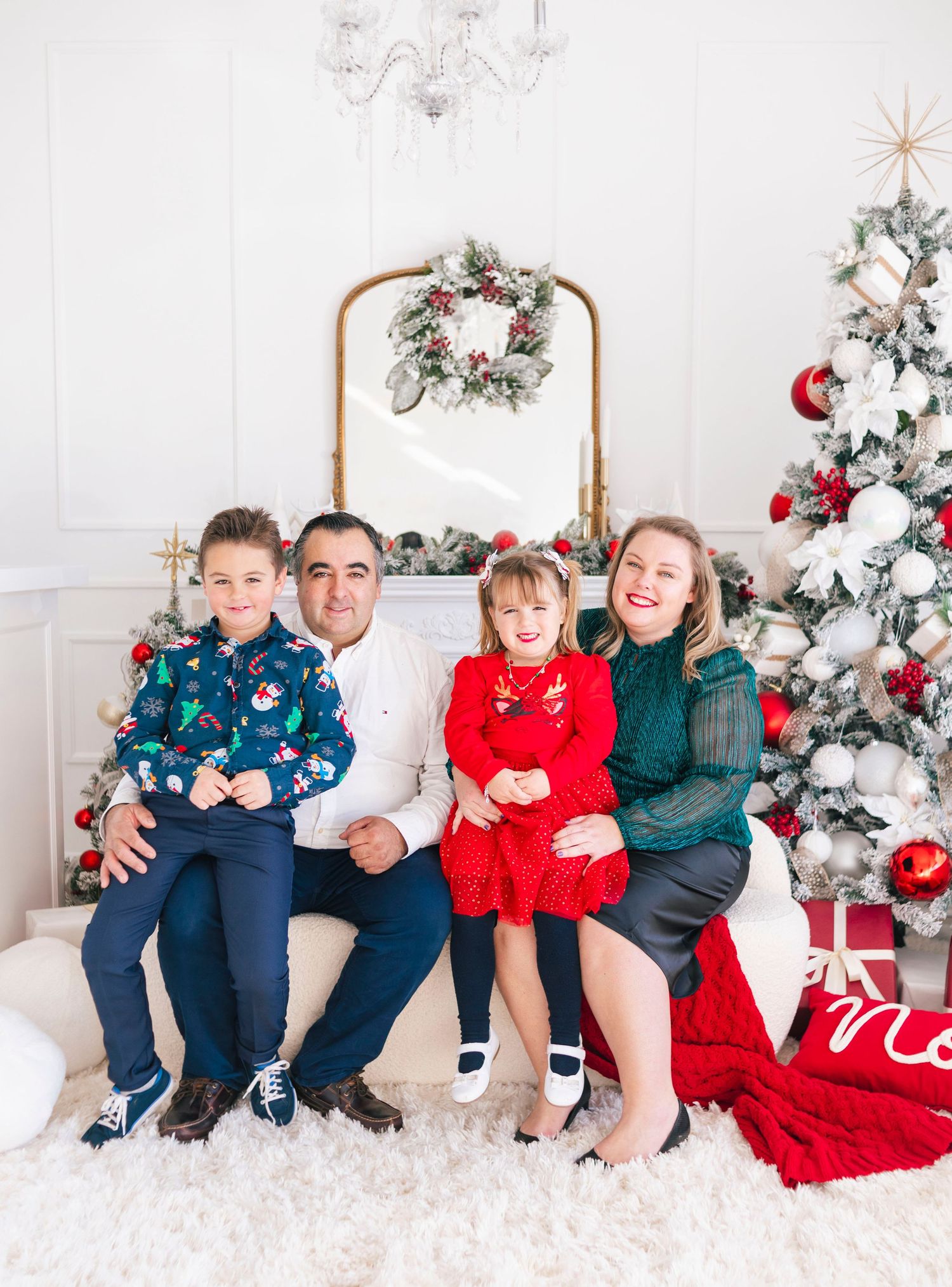 Family of four posing for a festive Christmas portrait with decorated trees, wreaths, and holiday decor in a bright white room.