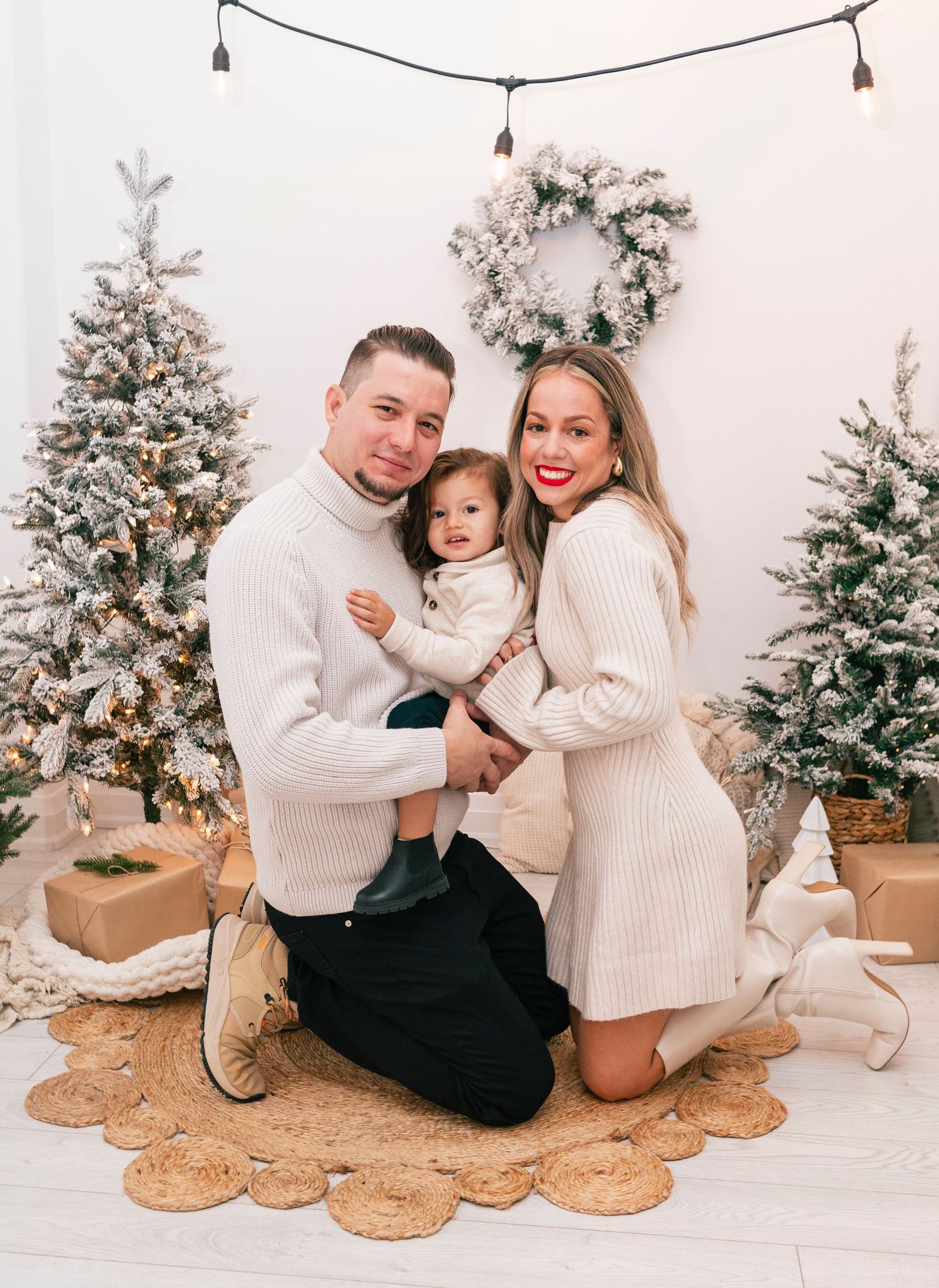 Family of three in matching cream sweaters posing together by flocked Christmas trees in a white studio.