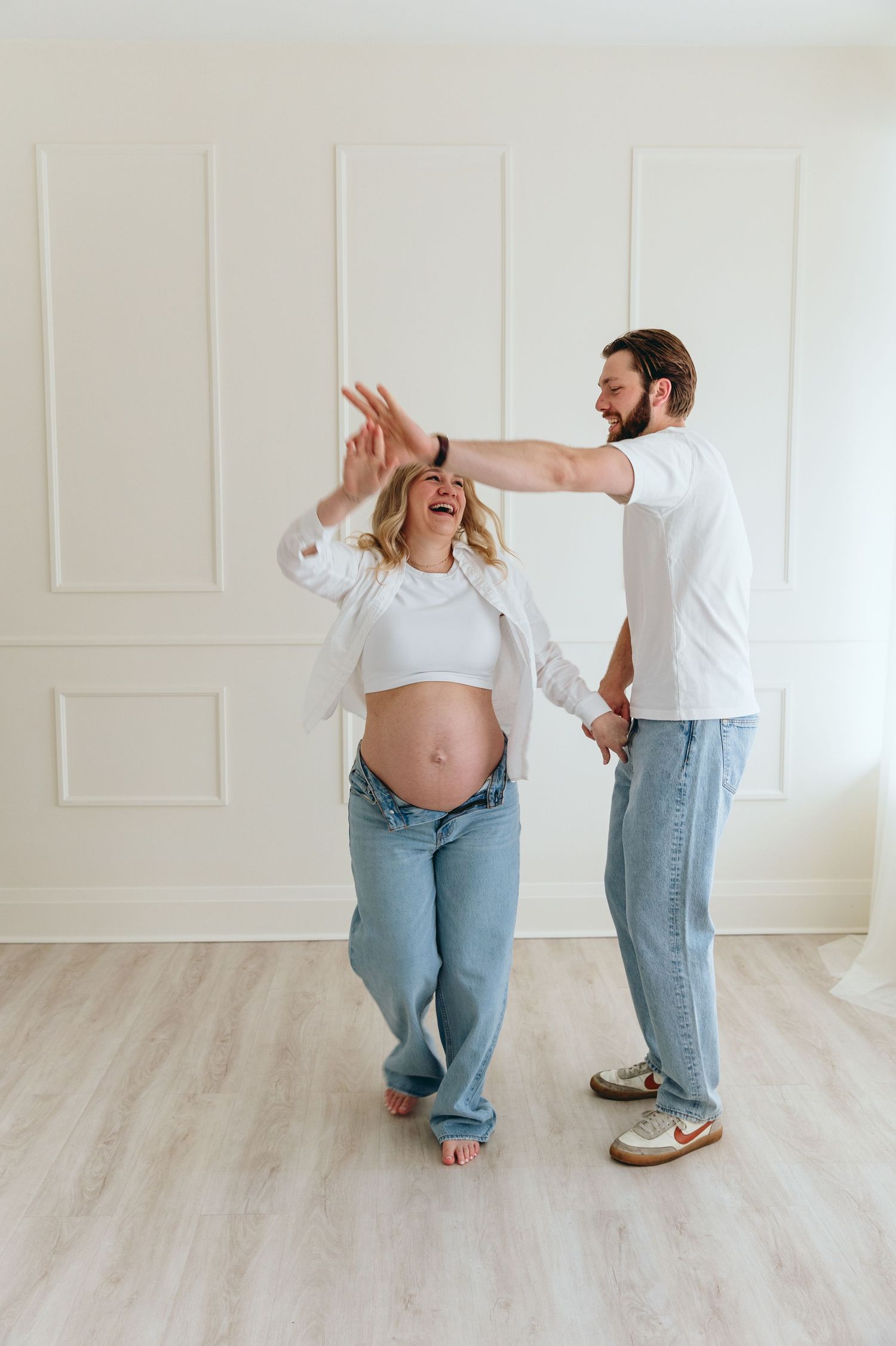 Playful maternity photoshoot showing couple dancing and laughing in bright white room.