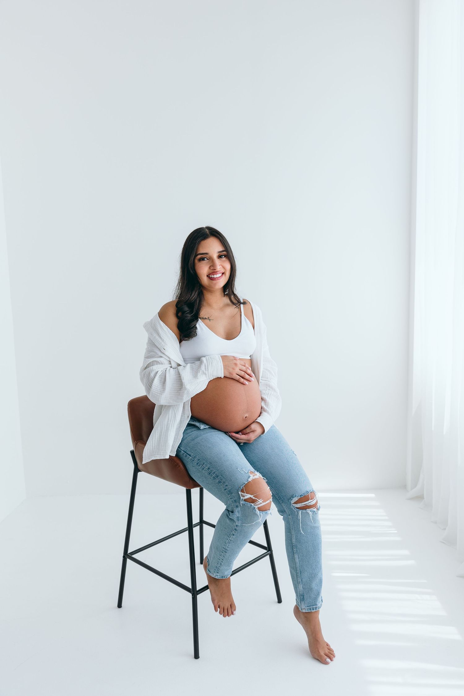 Pregnant woman in white top and ripped jeans sitting casually on chair in bright studio.