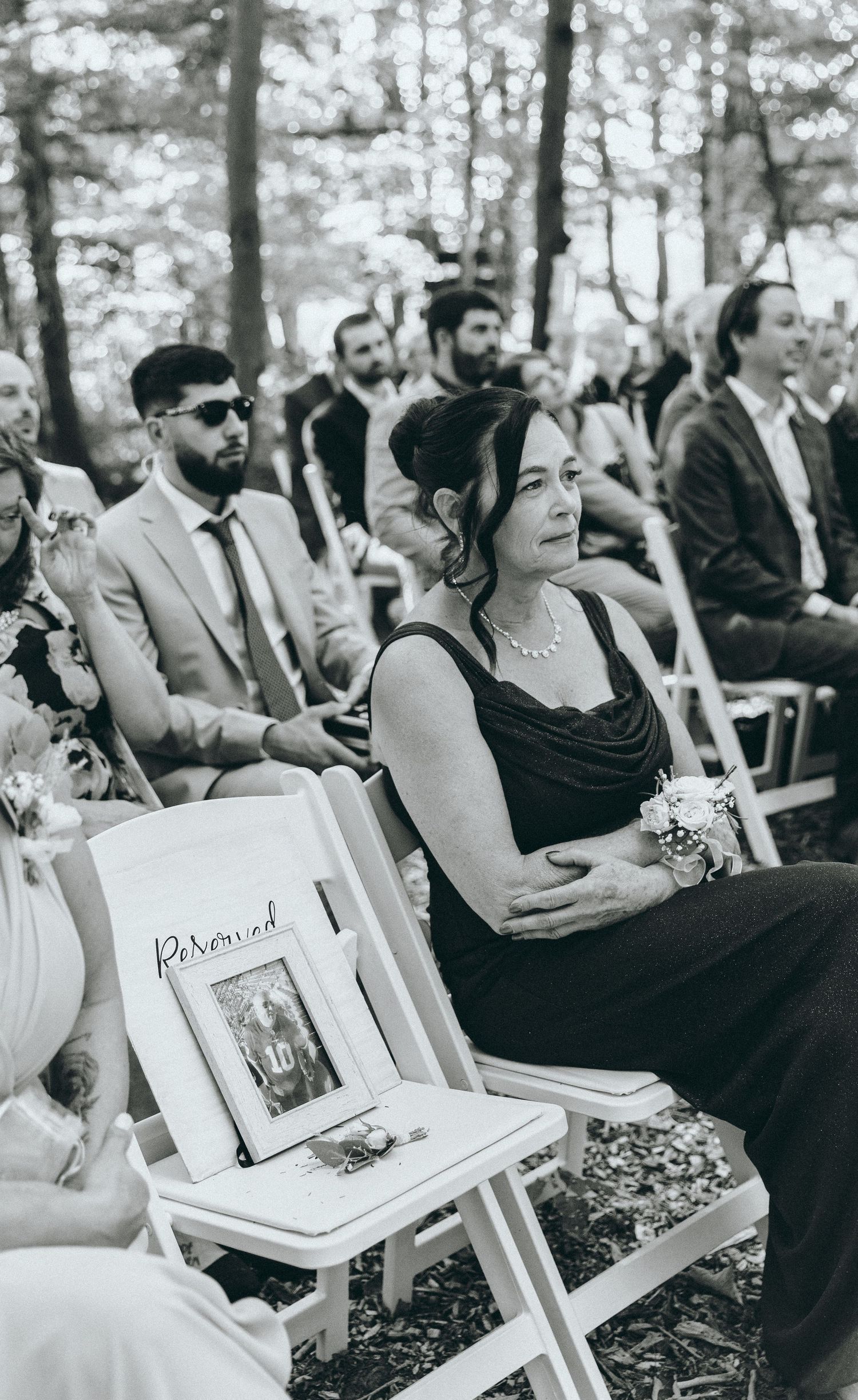 Black and white photo of wedding ceremony guests seated in white chairs among trees.