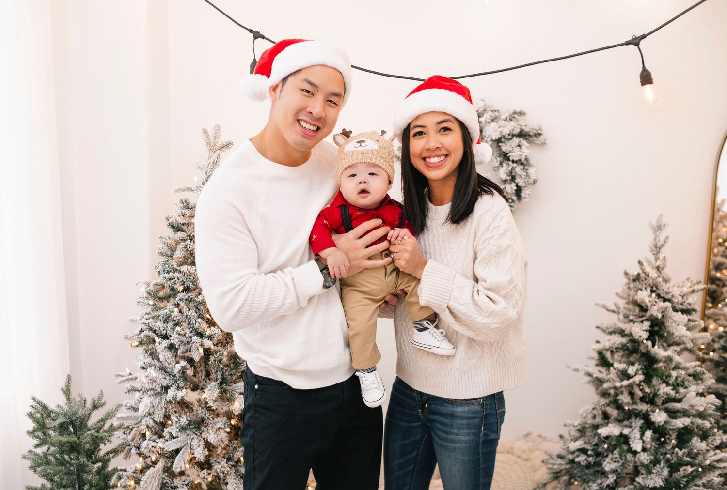 Family of three in Santa hats posing with snow-flocked Christmas trees for a festive holiday photo.