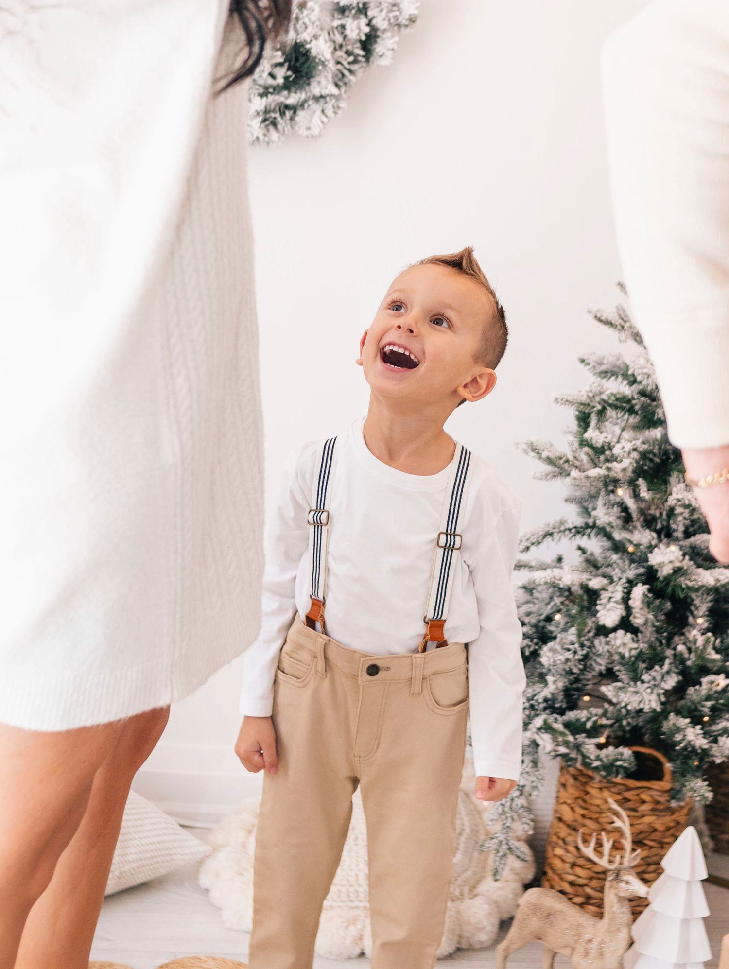 Happy young boy in suspenders and khakis laughs near decorated Christmas tree in bright white room.