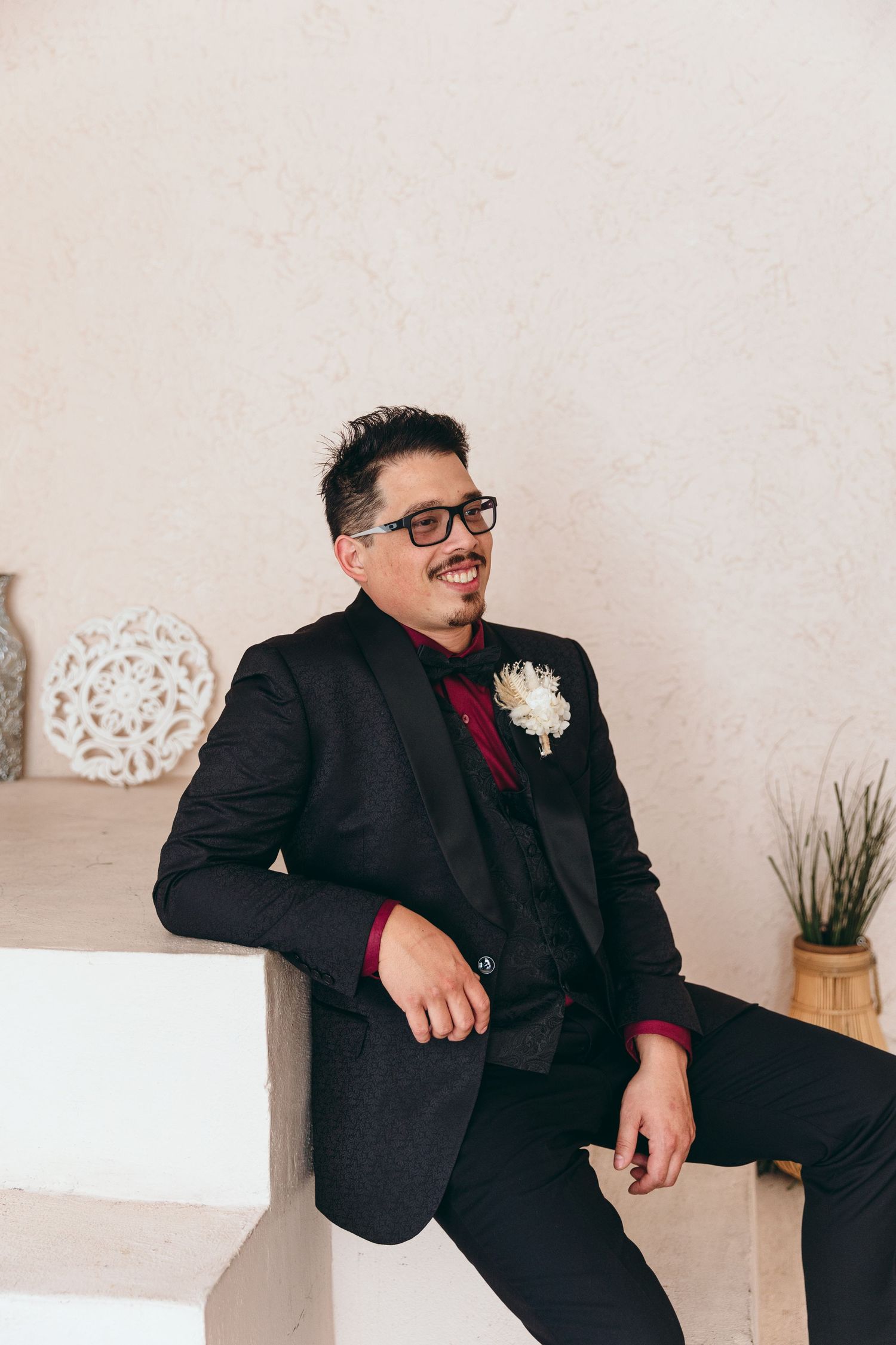 Groom in black suit and burgundy tie sits casually against light colored wall during wedding photoshoot.