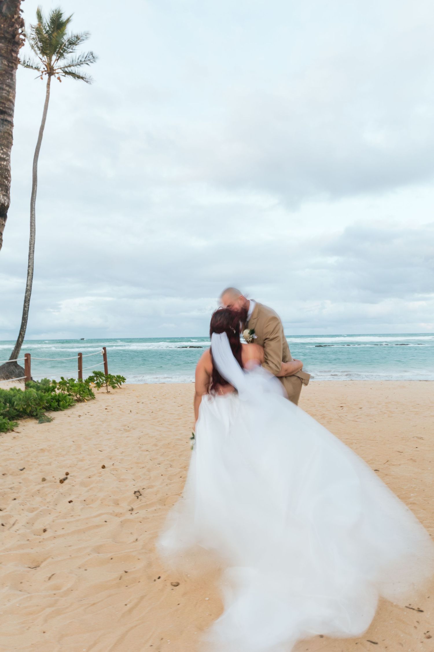 A romantic beach wedding photo with palm trees and ocean waves in the background.