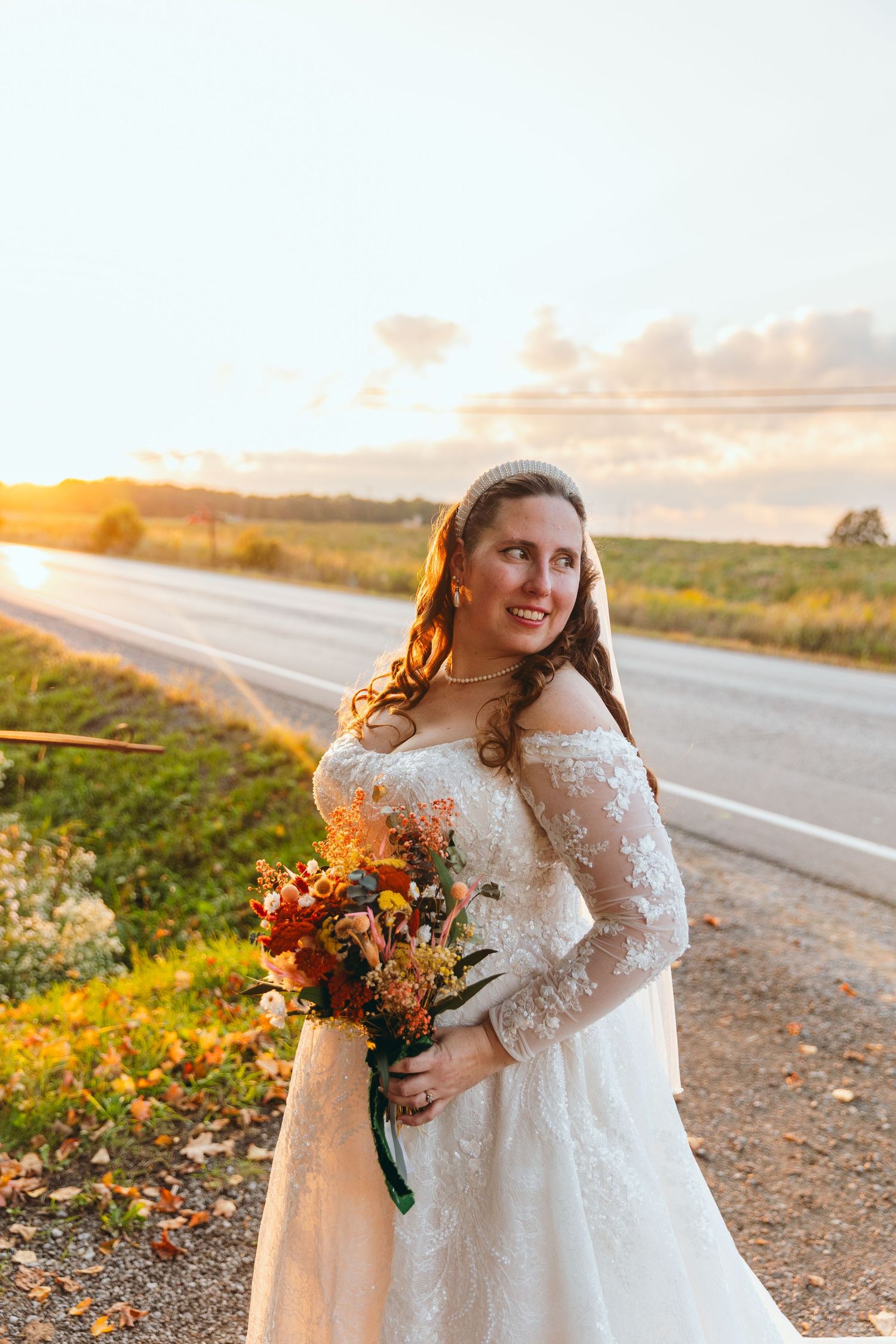 Bride in lace dress holds autumn bouquet during golden hour sunset.