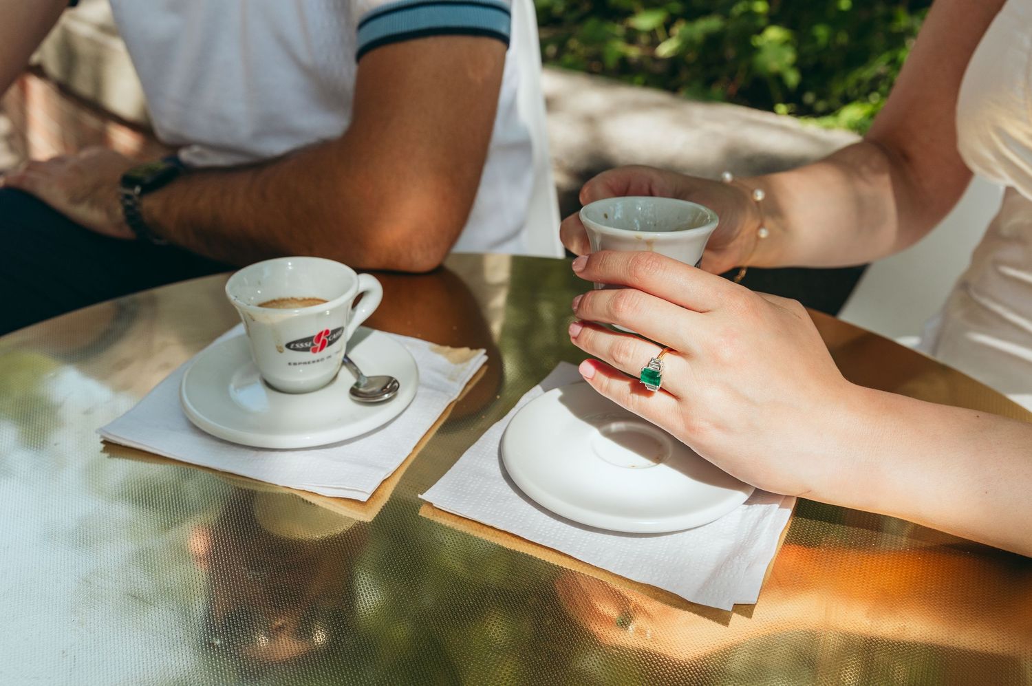 Coffee cups and hands shown on a metallic table surface in dappled sunlight.