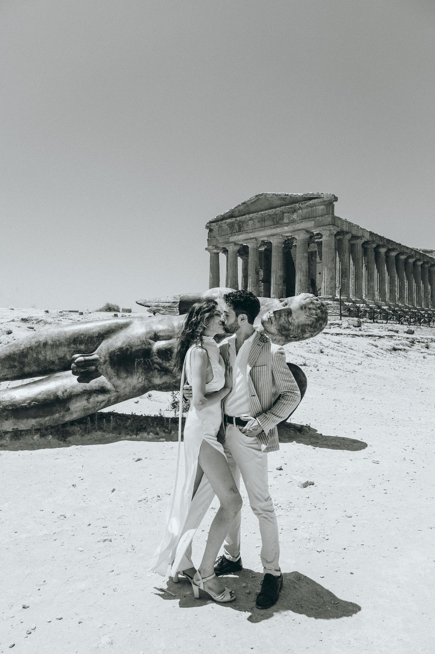 A couple in white outfits poses playfully near ancient Greek ruins in a black and white photograph.