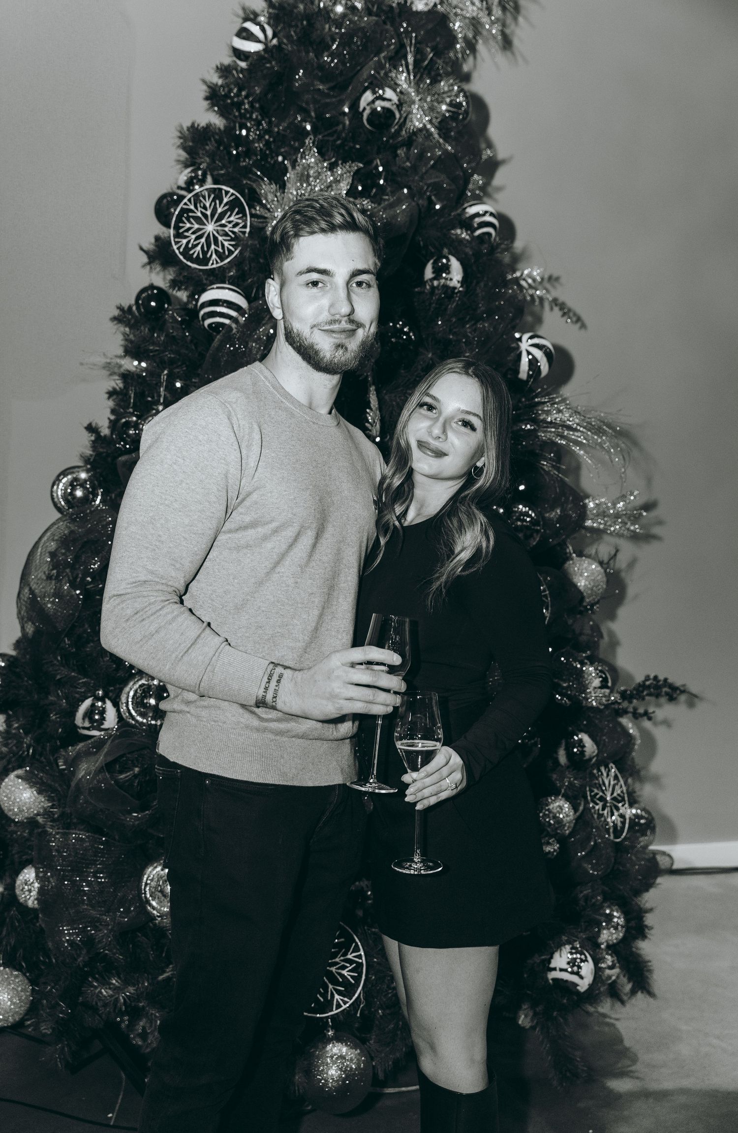 A couple poses together in formal attire in front of a decorated Christmas tree.