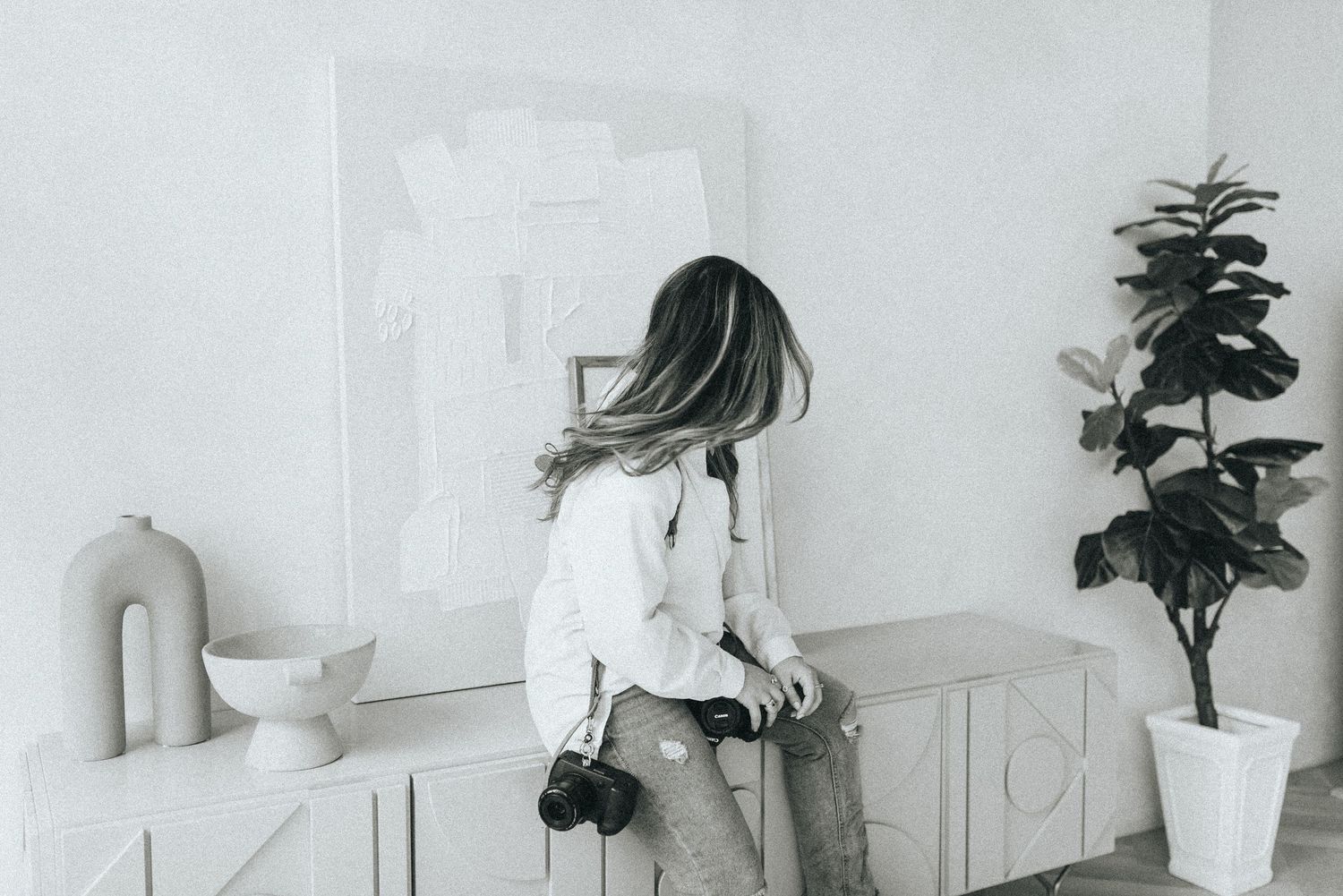 Figure in white sitting on counter against minimalist white wall with potted plant.
