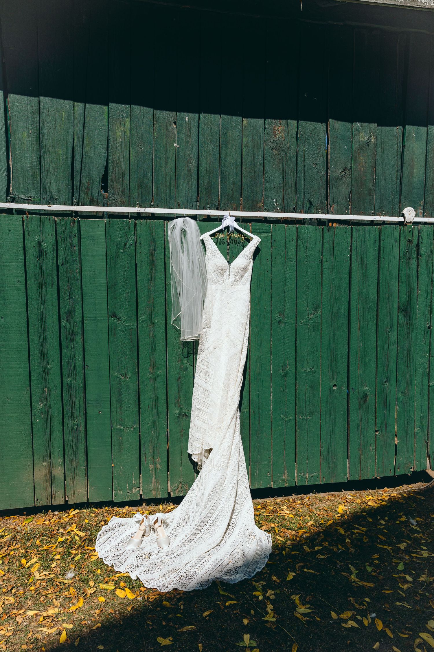 White wedding dress hanging on display against a green wooden fence with autumn leaves below.