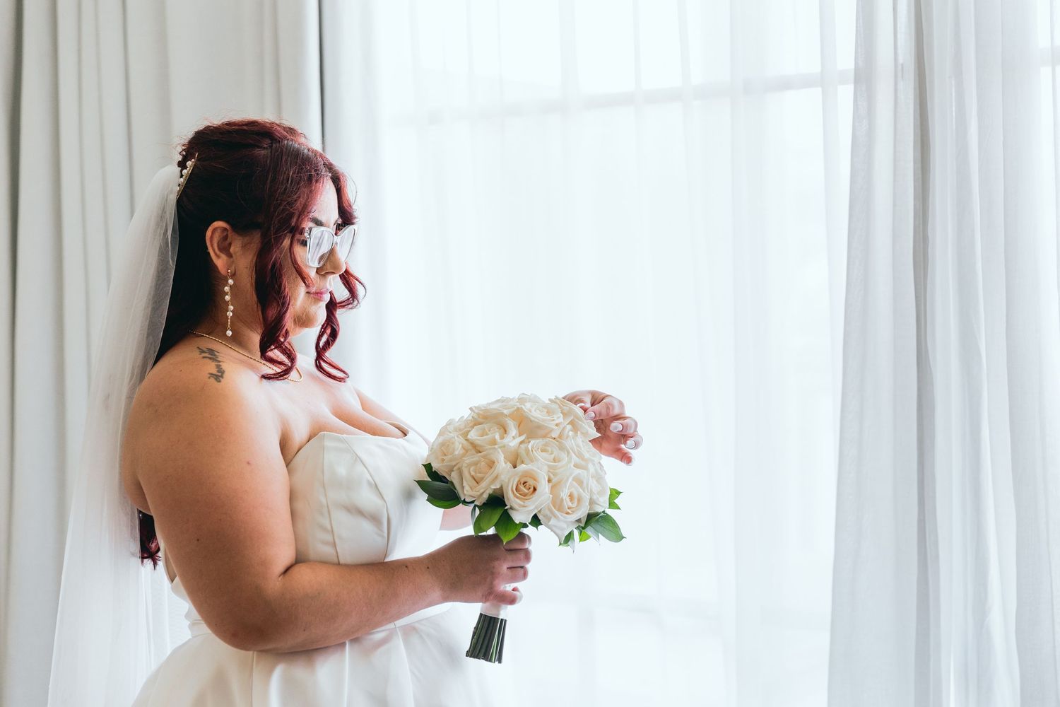 Bride in white dress holds a bouquet against bright window light.